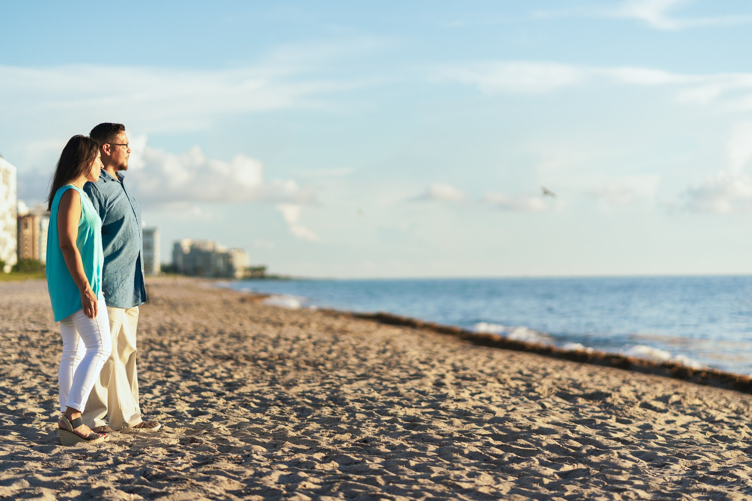 Couple standing on a sandy beach, gazing at the ocean, with a city skyline in the background under a partly cloudy sky.