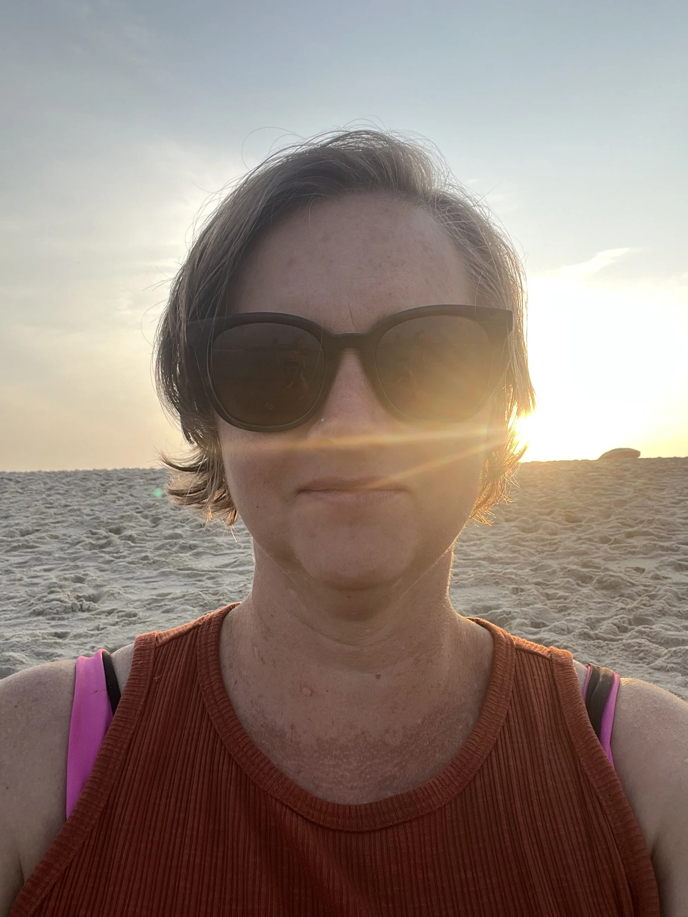 A woman with short brown hair wearing sunglasses and an orange tank top taking a selfie at the beach during sunset.