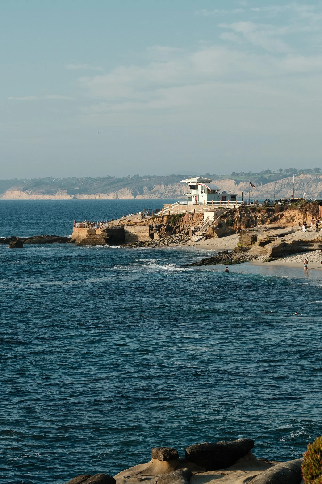 Coastal scene with rocky shoreline, beach, and a white building on a cliff overlooking the ocean, with a clear sky and distant hills in the background.