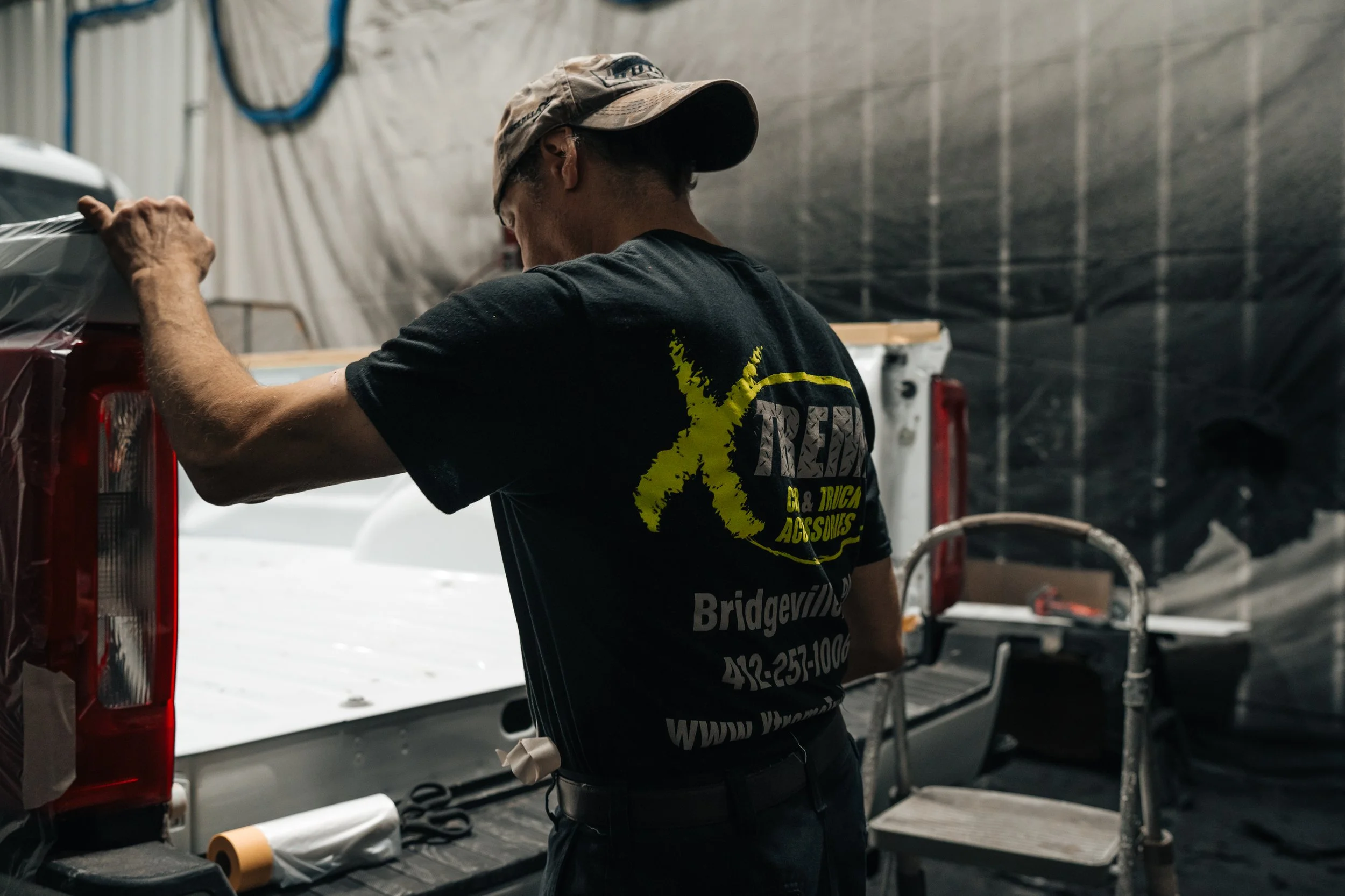 Technician at Xtreme Car & Truck Accessories in Bridgeville, PA applies protective masking inside a paint booth while preparing a white pickup truck for automotive refinishing work.