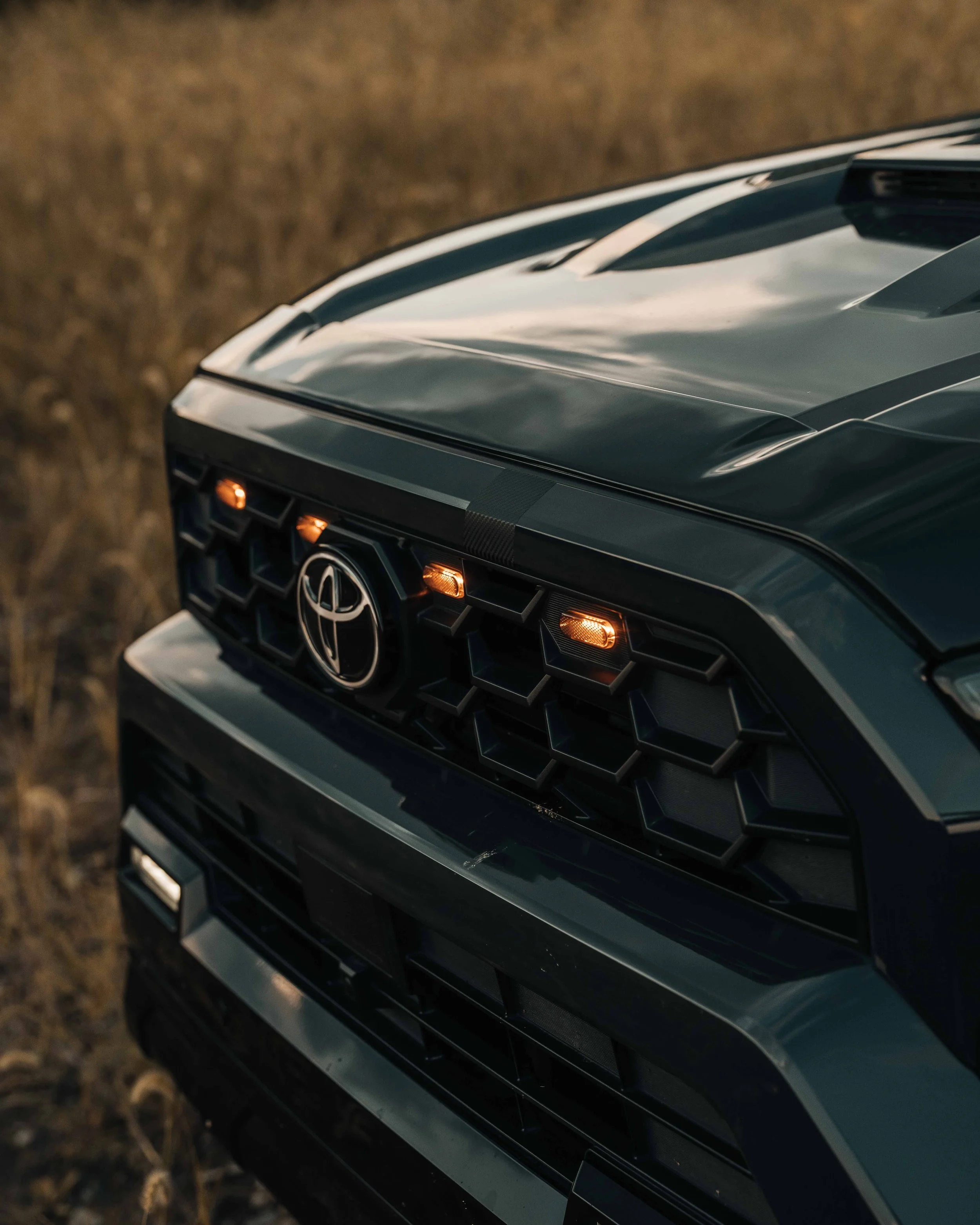 Front-end detail of Ian Jones' 2025 Toyota 4Runner TRD Sport in Underground gray showing four amber raptor-style grille lights lit around the Toyota emblem, the hood scoop line, and the honeycomb TRD Sport grille in low evening light.