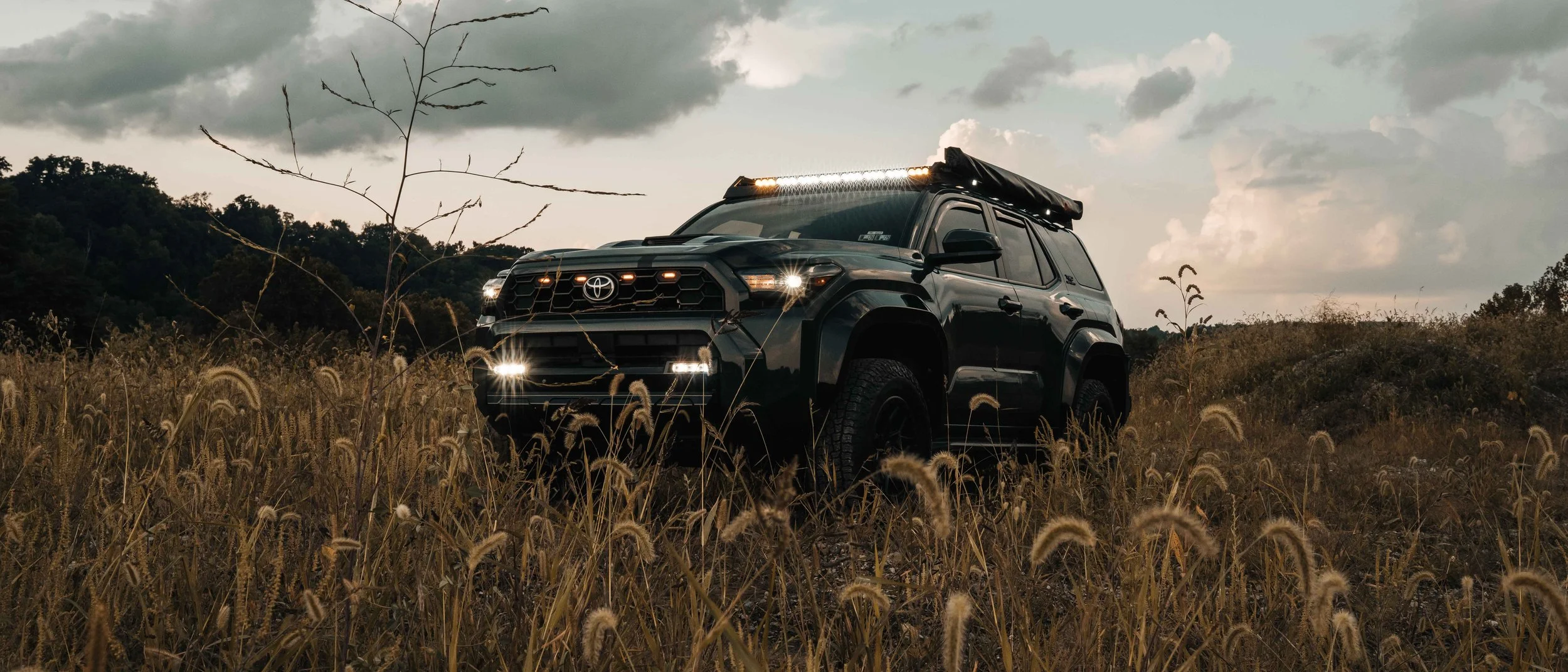 Wide front three-quarter view of Ian Jones' 2025 Toyota 4Runner TRD Sport in Underground gray standing in tall fall grass with the Cali Raised LED roof rack light bar, grille lights, and bumper lights on against a cloudy Western Pennsylvania sky.