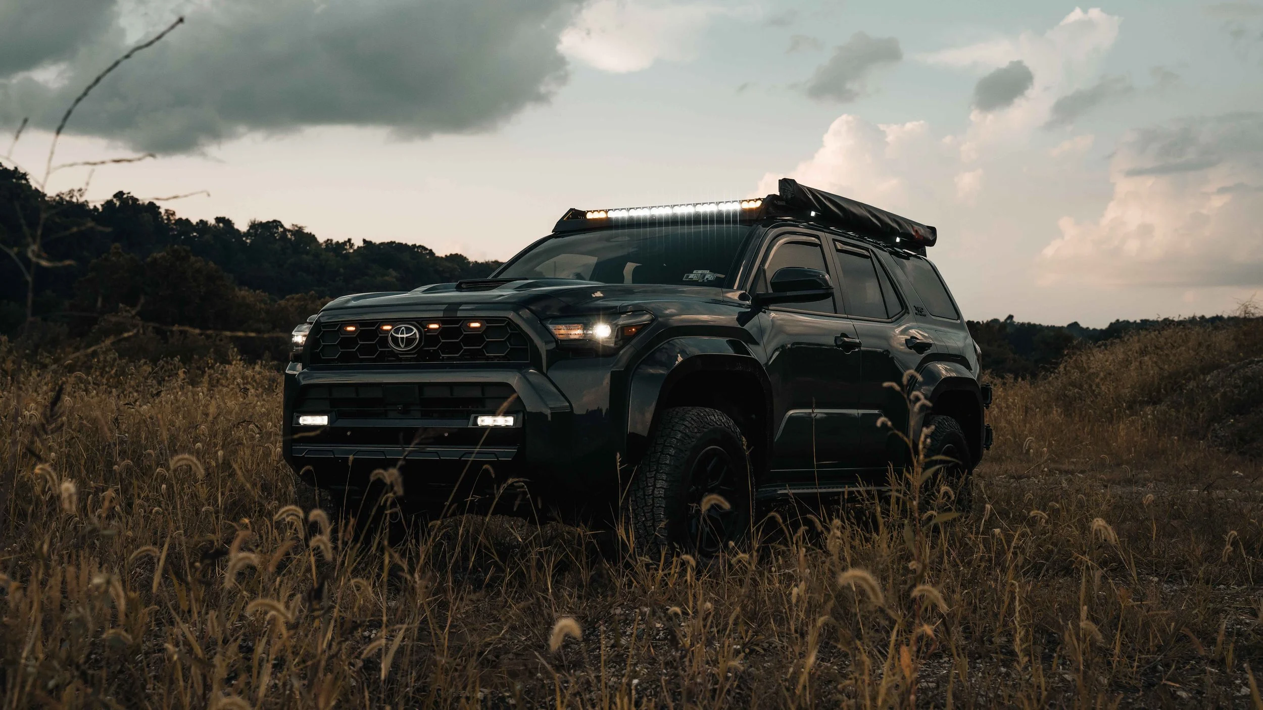 Front three-quarter view of Ian Jones' 2025 Toyota 4Runner TRD Sport in a dusk field with the Cali Raised LED roof rack light bar, amber grille lights, and lower LED bar all illuminated, showing the full 6th gen 4Runner lighting setup and stowed awning on the rack.