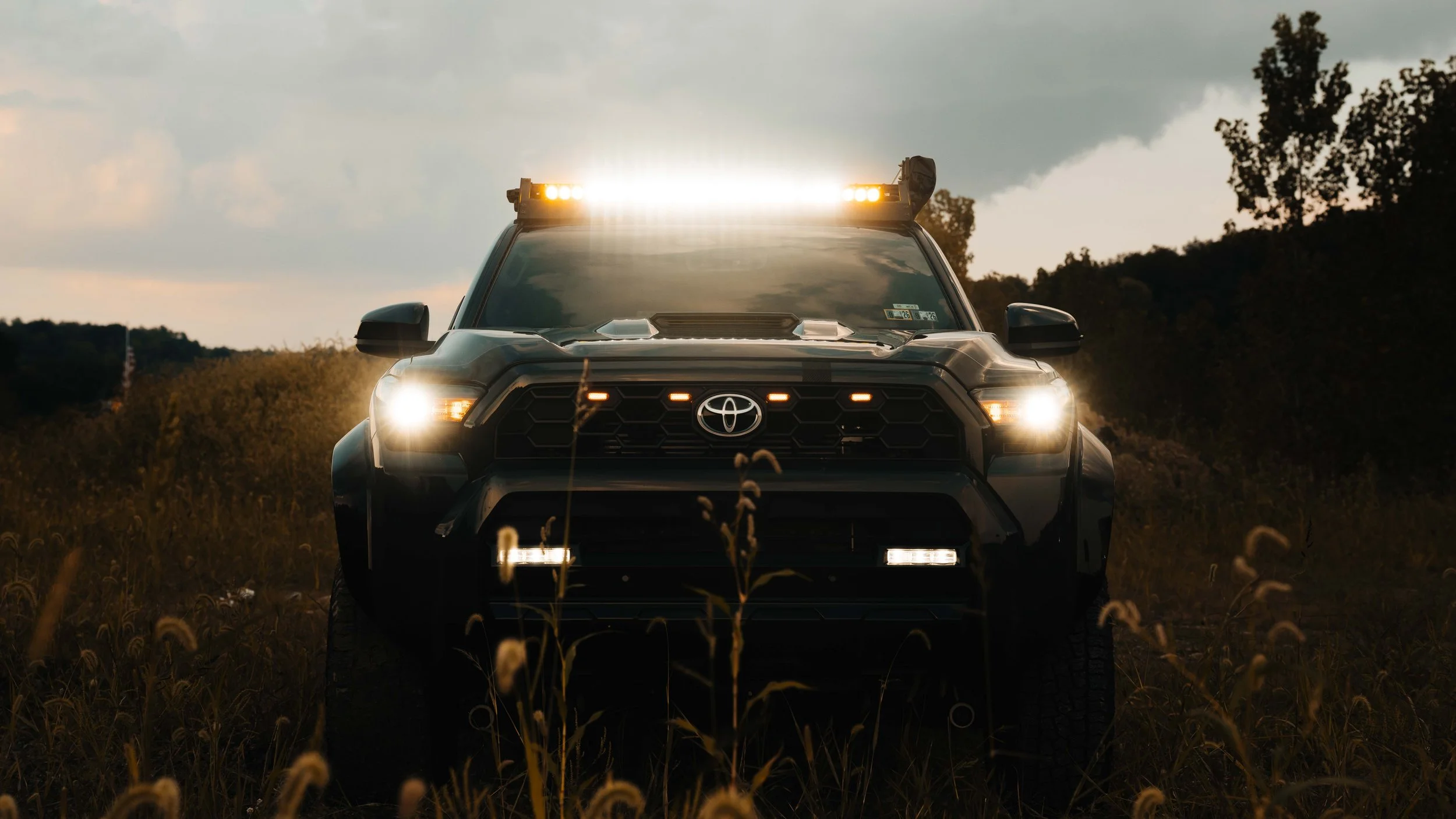 Head-on front view of Ian Jones' 2025 Toyota 4Runner TRD Sport with the Cali Raised LED 43 inch dual function light bar running white center optics and amber ends, plus illuminated grille lights, LED headlights, and lower bumper light bars in a field at dusk.