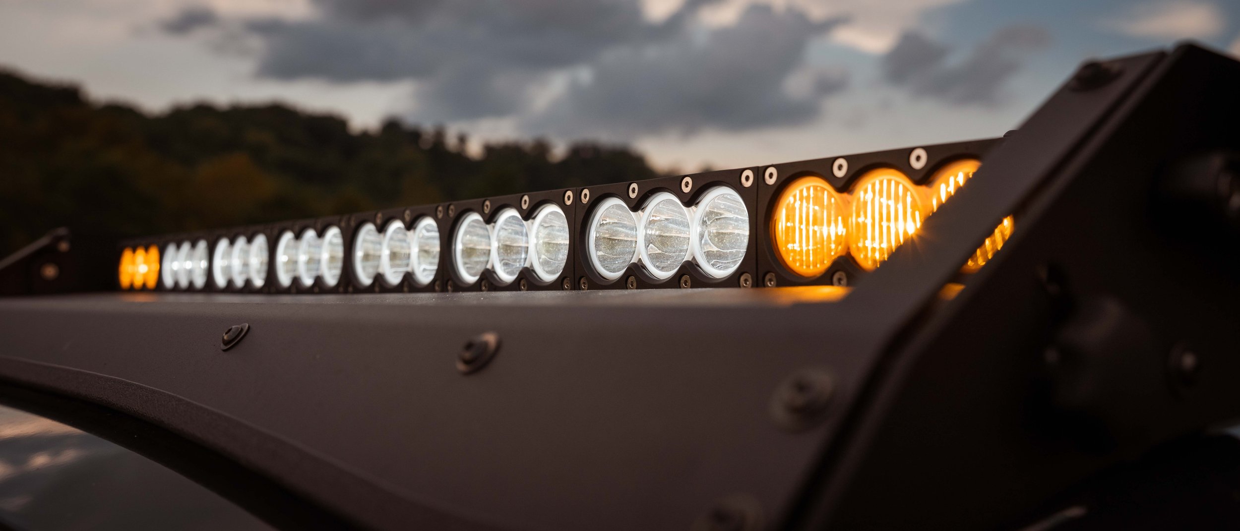 Close-up of the Cali Raised LED 43 inch dual function light bar mounted on the Premium Roof Rack of Ian Jones' 2025 Toyota 4Runner TRD Sport, showing white forward optics and amber outboard optics lit at dusk above the black powder-coated adjustable wind deflector.
