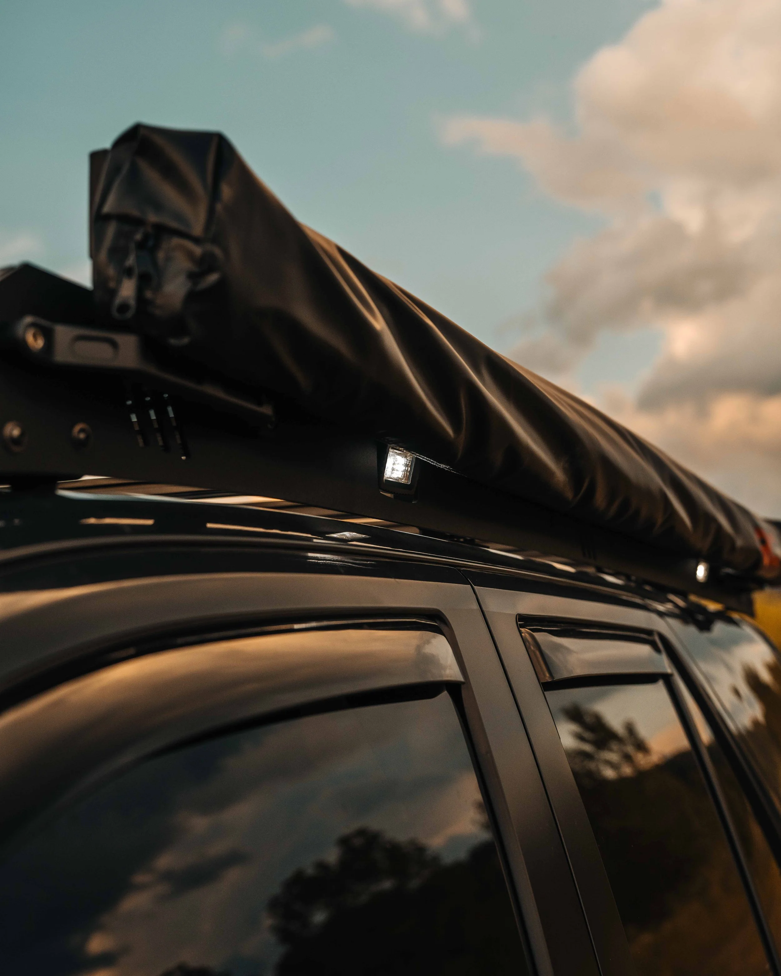 Close-up of the Rough Country retractable awning stowed in its black cover along the driver side of the Cali Raised LED roof rack on a 2025 Toyota 4Runner TRD Sport, showing the mounting bracket, a side LED pod, and window deflectors below.