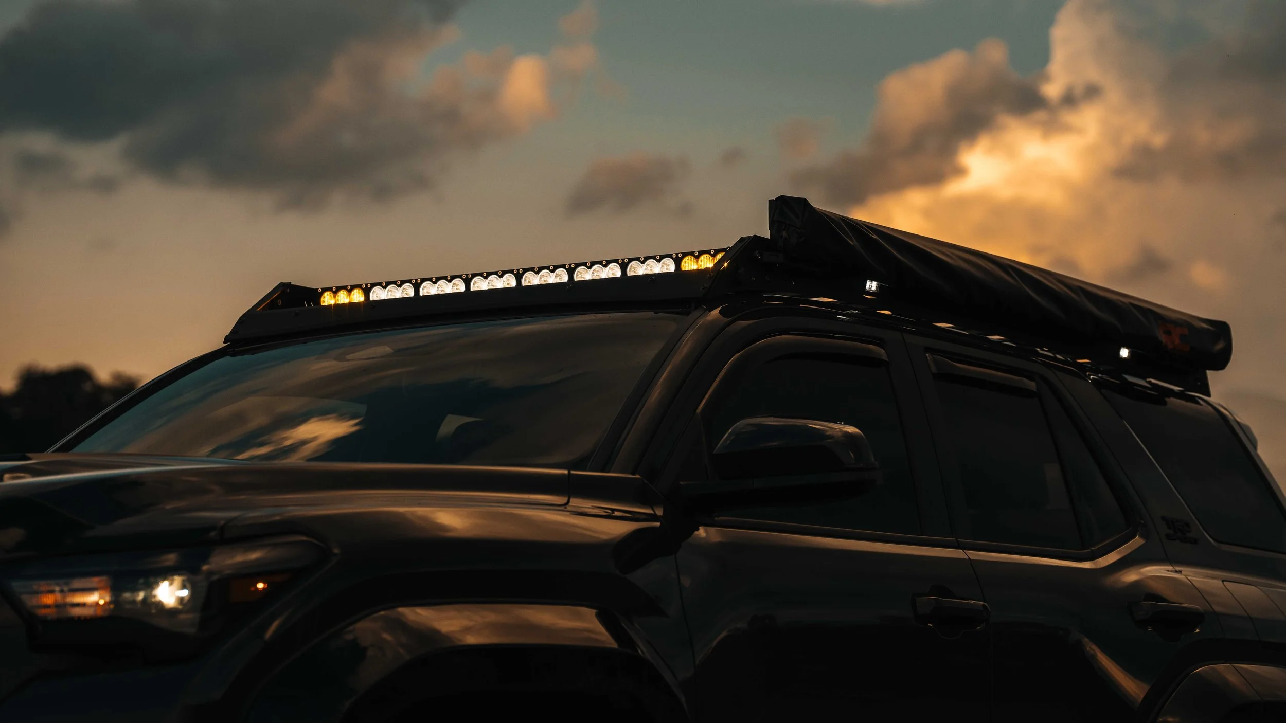 Roofline detail of Ian Jones' 2025 Toyota 4Runner TRD Sport at sunset with the Cali Raised LED 43 inch light bar lit across the front of the Premium Roof Rack, the Rough Country awning stowed along the passenger side, and window deflectors visible below the rack.