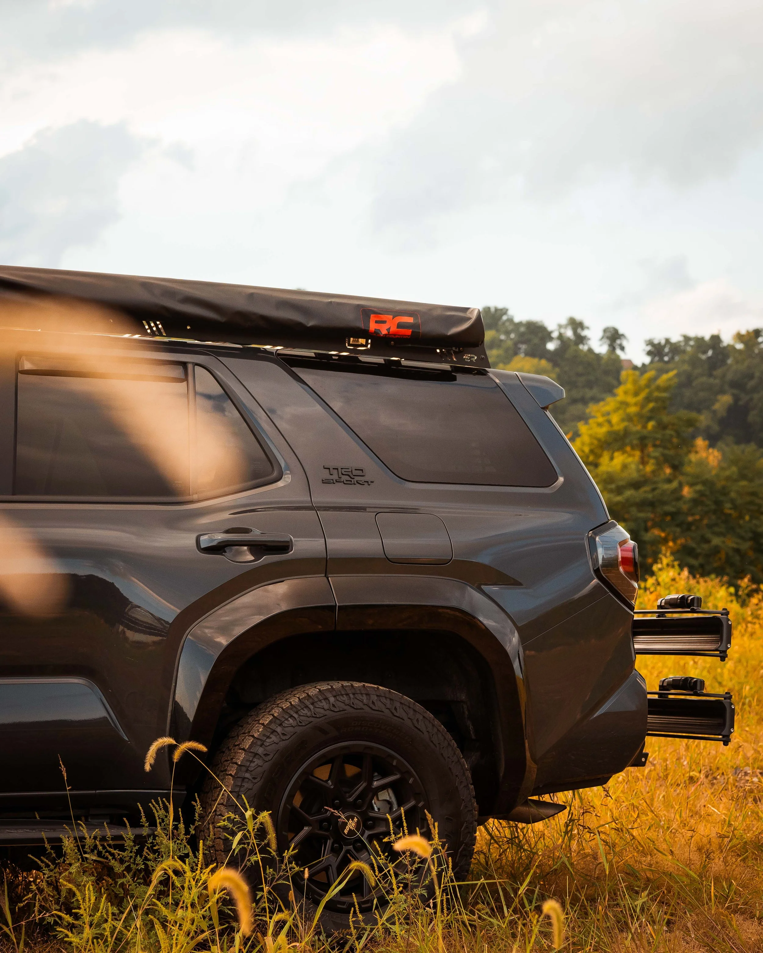 Rear three-quarter view of Ian Jones' 2025 Toyota 4Runner TRD Sport in golden evening light showing the Rough Country retractable awning stowed on the roof rack, the TRD Sport quarter-panel badge, gloss black fender flares, and a rear hitch-mounted rack.