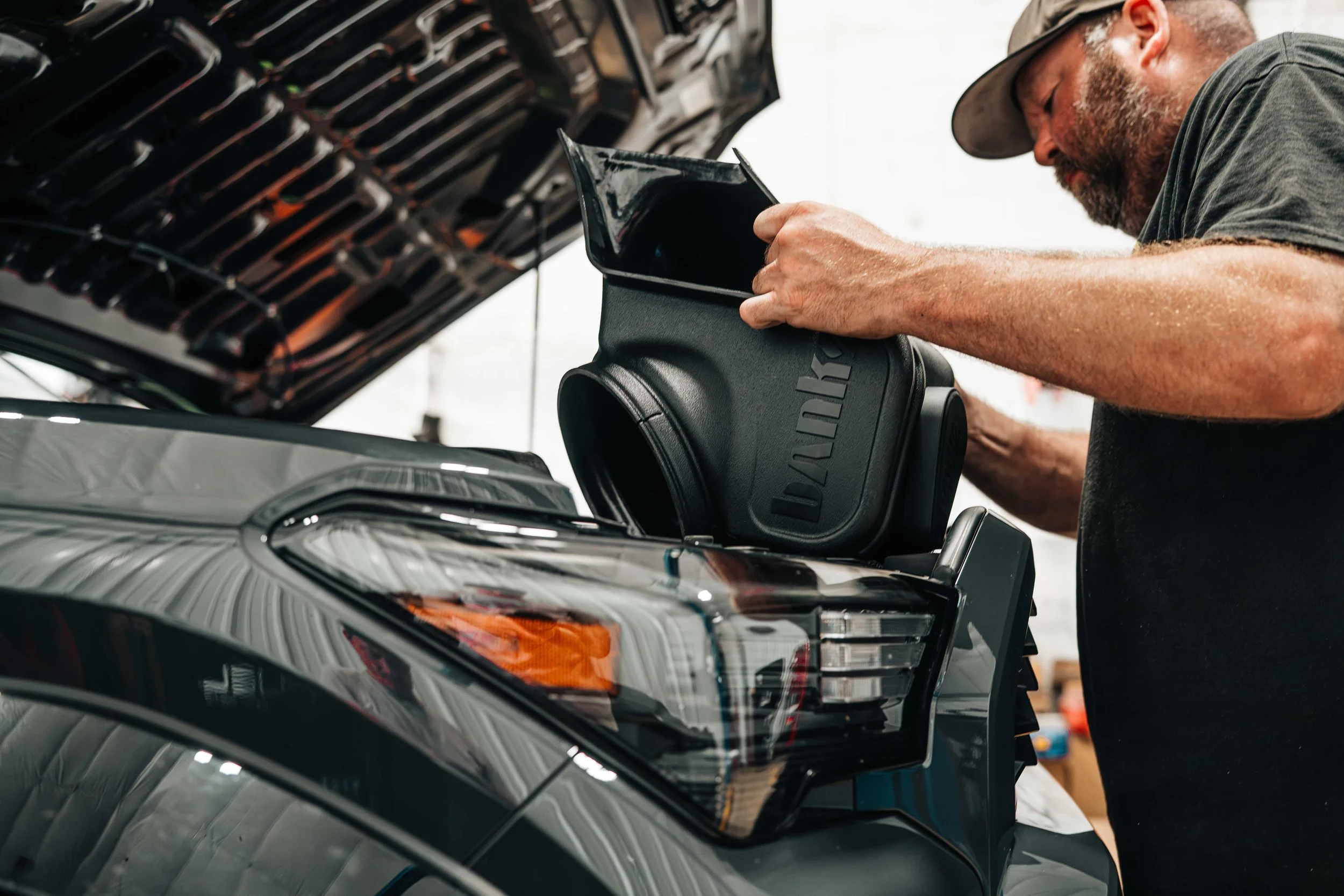 Banks Ram-Air intake housing being lifted into place over the engine bay of a 2025 Toyota 4Runner TRD Sport during installation, framed past the truck's LED headlight, amber marker, and Underground gray front fender.