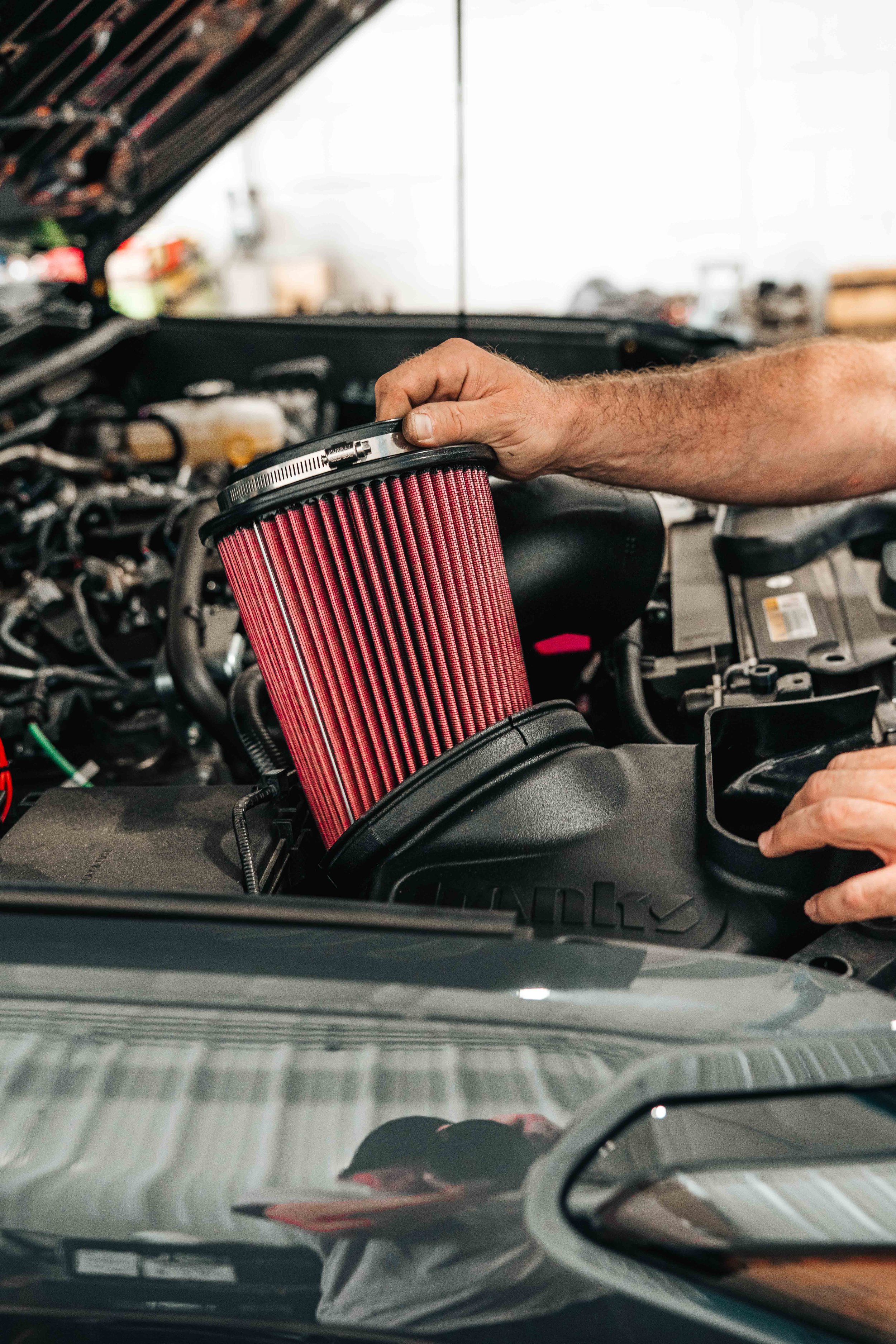 Red conical Banks Ram-Air filter being lowered into the intake housing on a 2025 Toyota 4Runner TRD Sport during installation, with the clamp and housing lip visible and the raised hood reflecting the shop above.