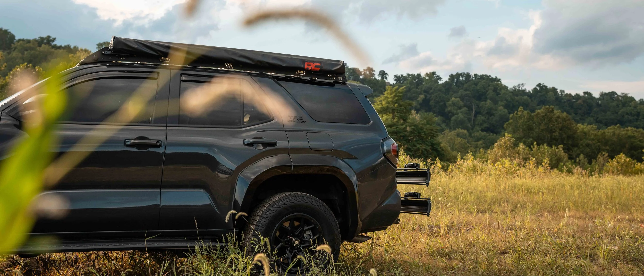Rear three-quarter view of Ian Jones' 2025 Toyota 4Runner TRD Sport in Underground gray parked at the edge of a field, showing the roof rack with stowed Rough Country awning, rear hitch rack, wide fender flares, and all-terrain tires.