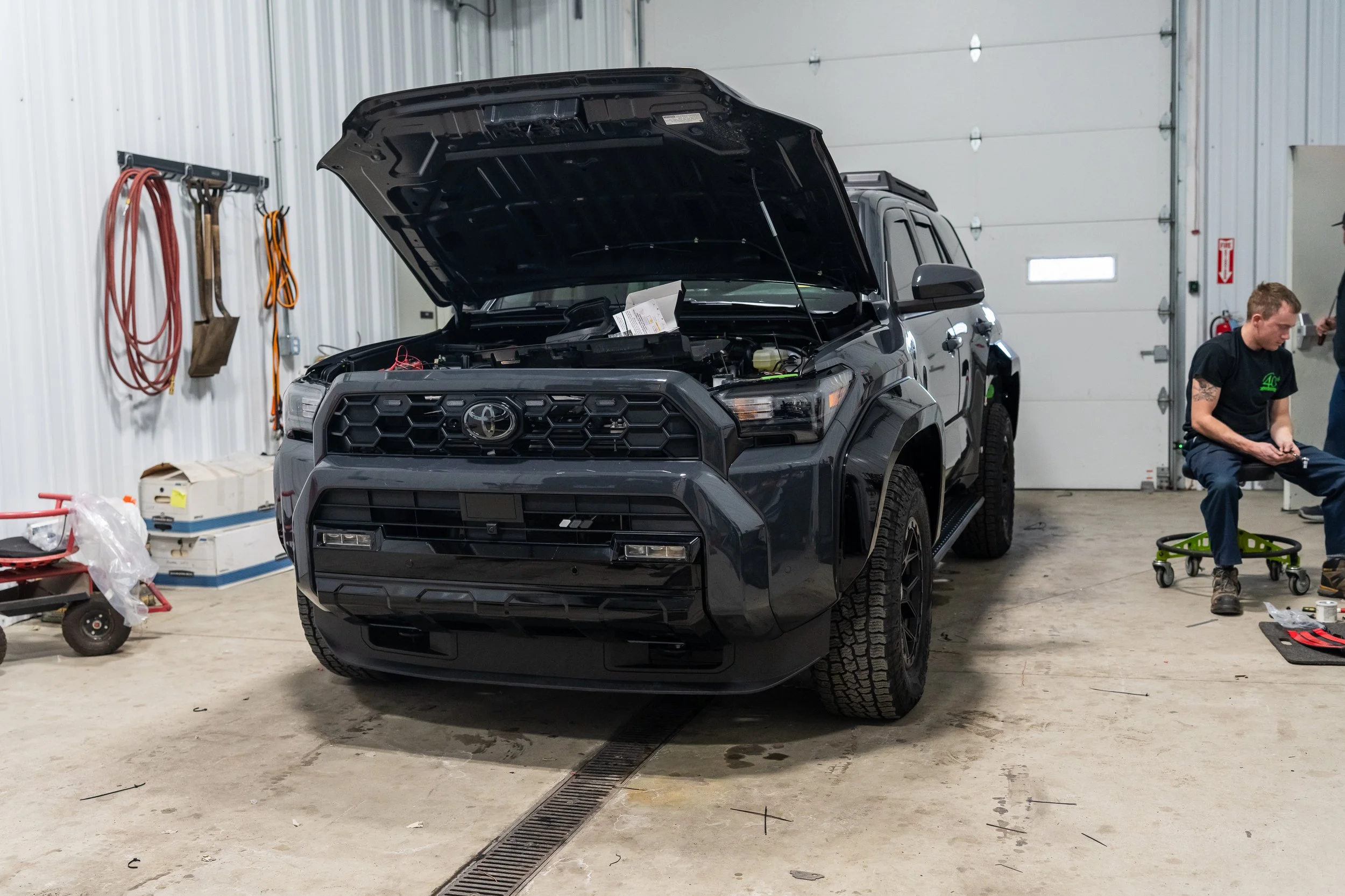 2025 Toyota 4Runner TRD Sport with hood open in shop, featuring Rocktrix 17x8.5 ET+25 wheels, 265/70R17 Cooper Discoverer AT Trail tires, and Rough Country gloss black fender flares.