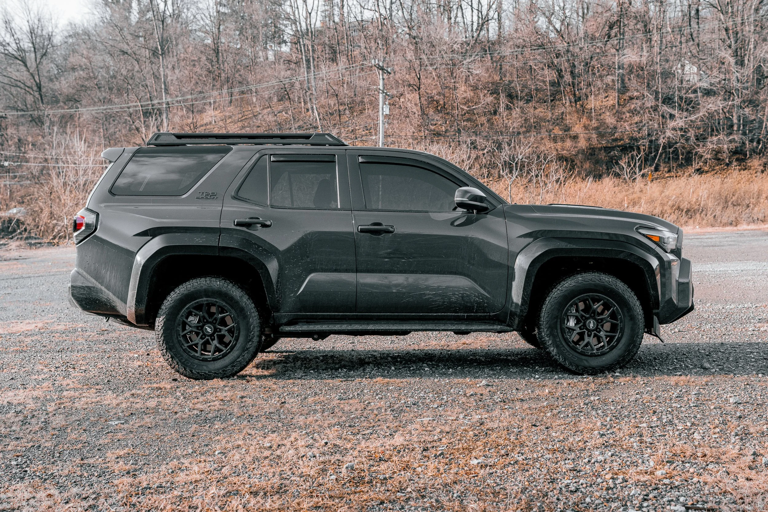 Side profile of a 2025 Toyota 4Runner TRD Sport in dark gray with black wheels and all-terrain tires, parked on a gravel lot in winter.