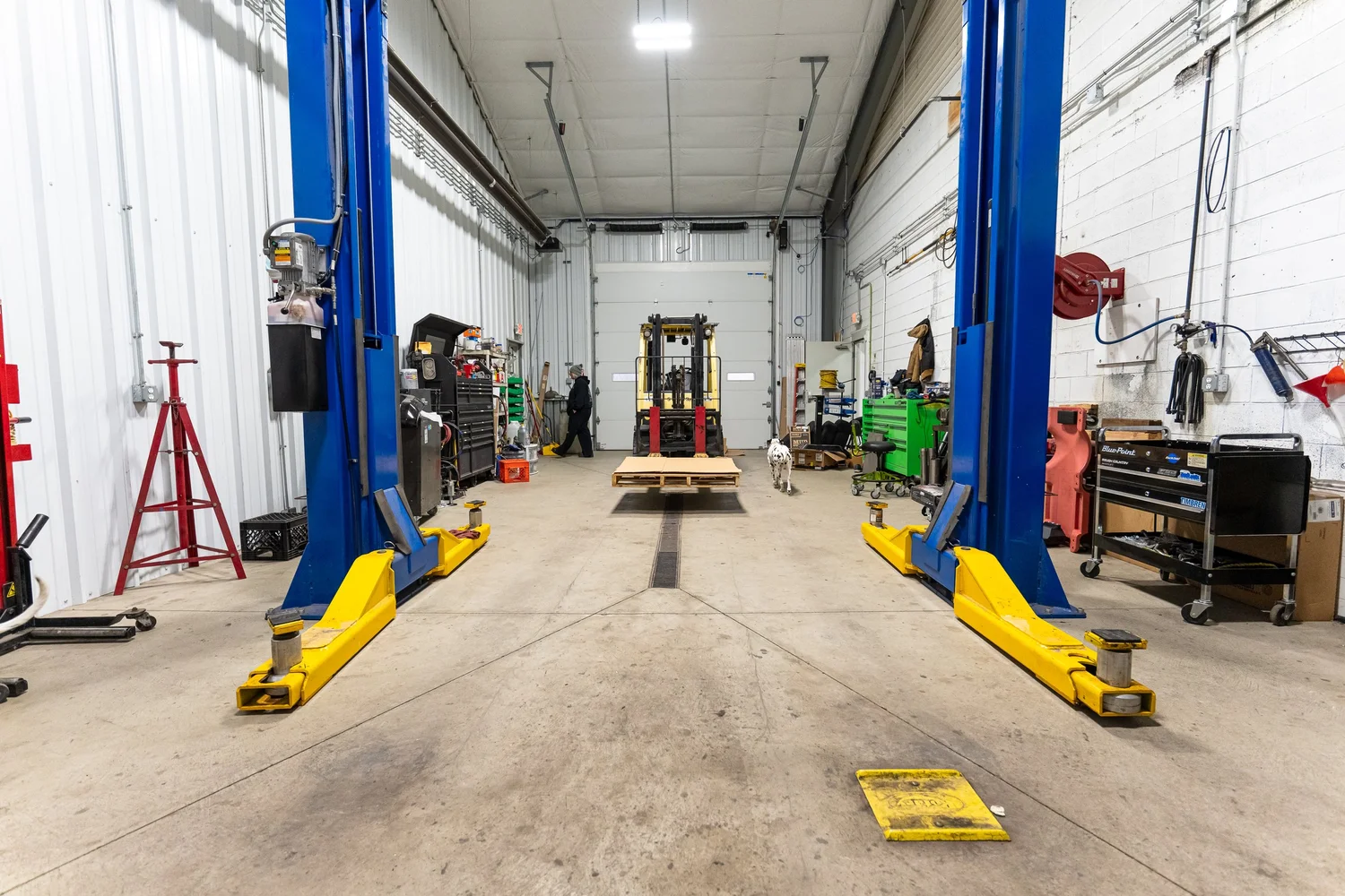 Wide view of a Pittsburgh automotive shop install bay with blue two-post car lift, forklift, and mechanic working in the background