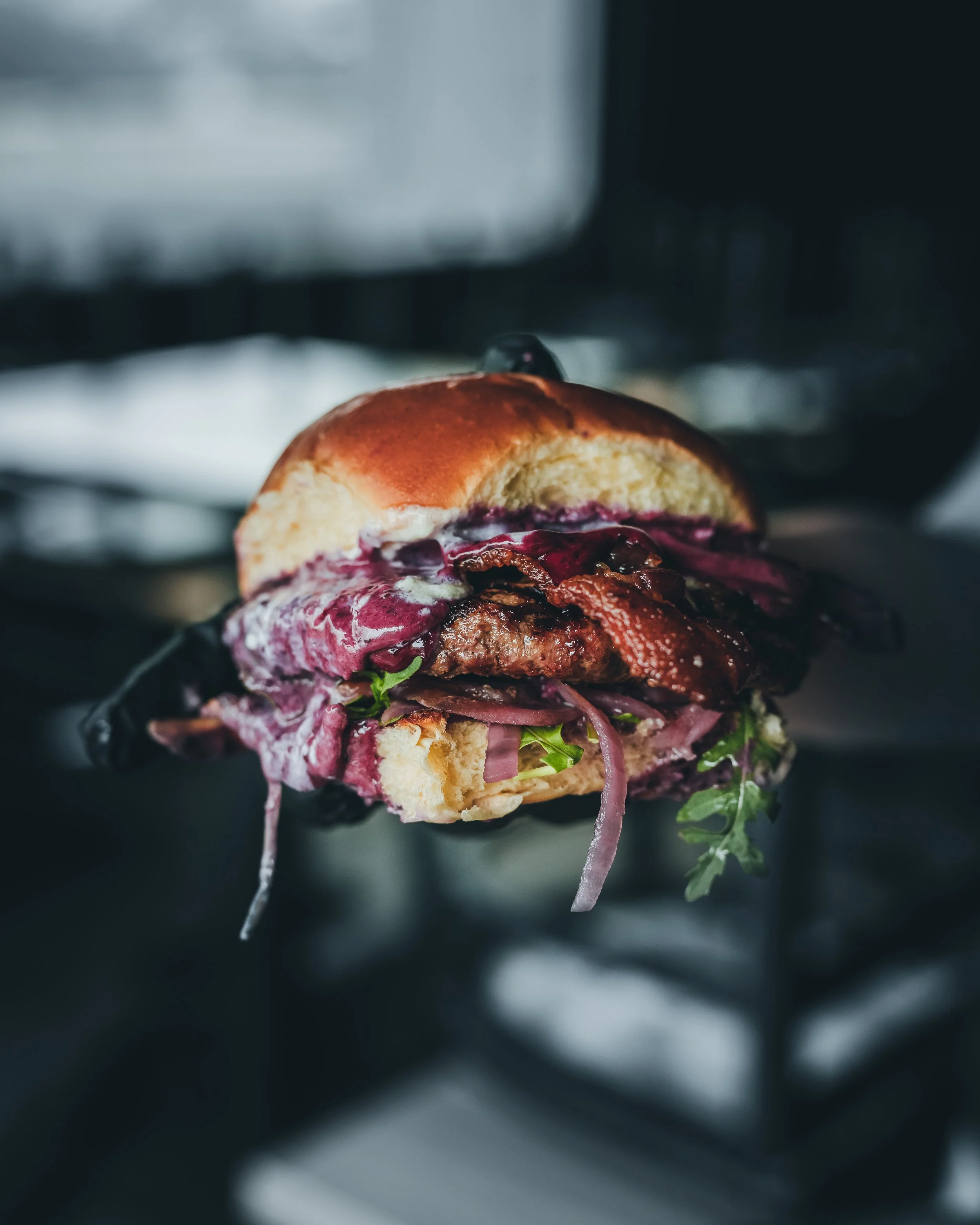 Dramatic restaurant burger photography with bacon, onions, greens, and shallow depth of field by Ian Jones