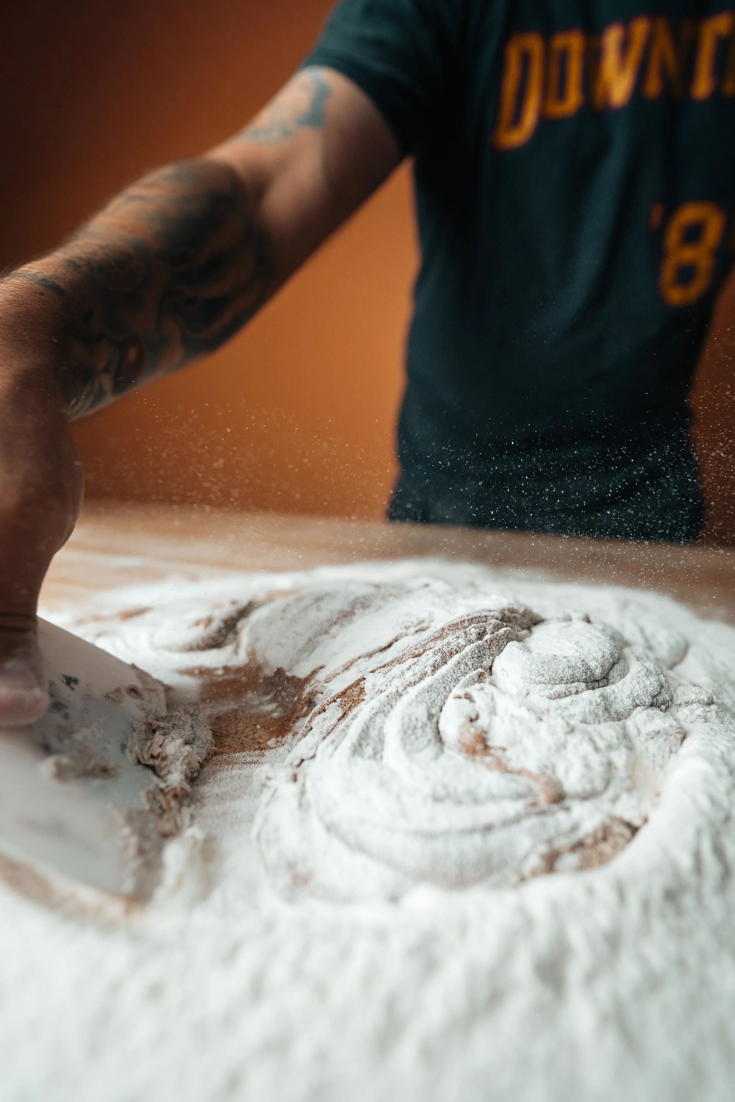Action shot of a baker using a bench scraper on flour-covered dough with flour particles suspended in the air, captured on Sony a1 with the Tamron 16-30mm f/2.8 G2 by Pittsburgh photographer Ian Jones.