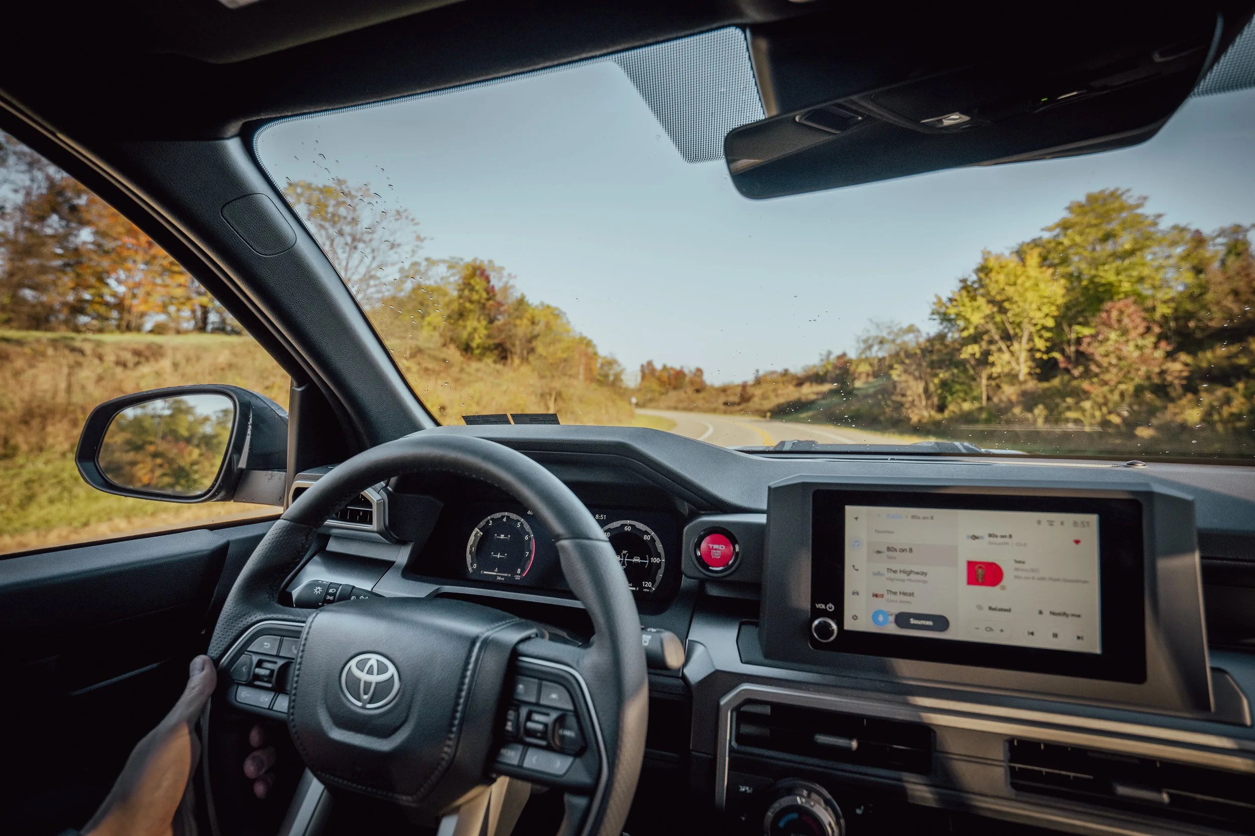 Driver’s-eye view inside a 2025 Toyota 4Runner TRD Sport: Toyota-badged steering wheel, gauges, red TRD start button, and center touchscreen visible while cruising a curving rural road lined with early fall trees; light raindrops on the windshield.”