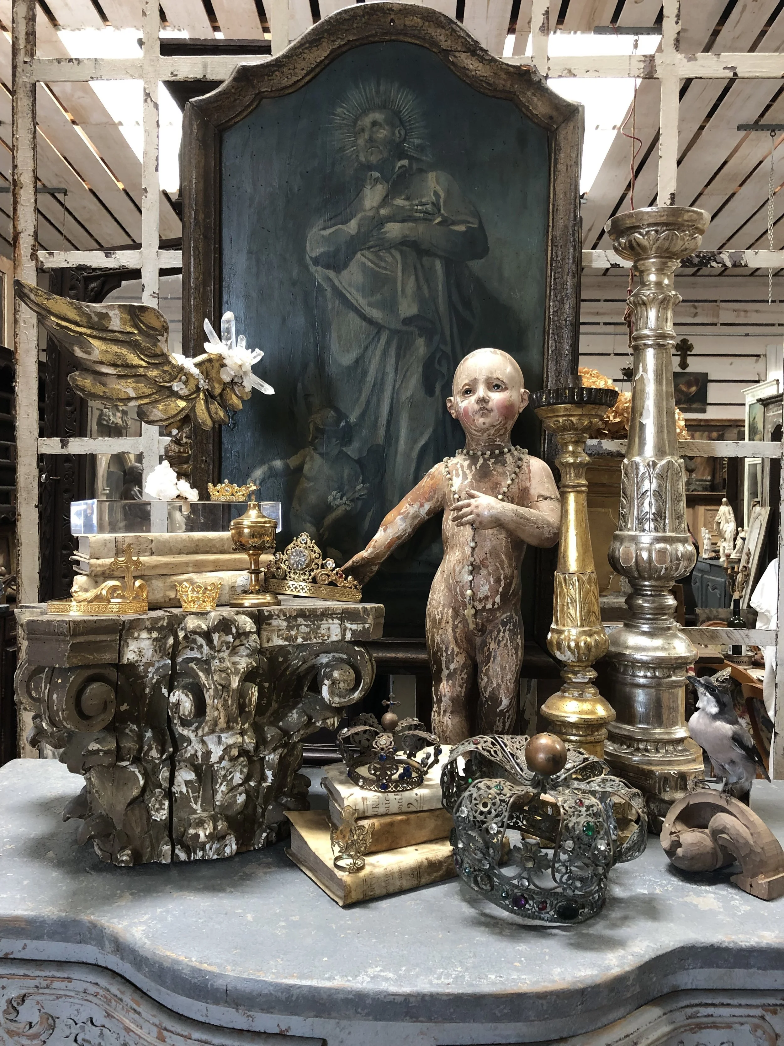 An 18th-century Italian Christ Child is surrounded by treasures found on the latest buying trip. Antique crowns, altar sticks, vellum books, and an 18th- century angel’s wing mounted on a modern base come together to tell a story.