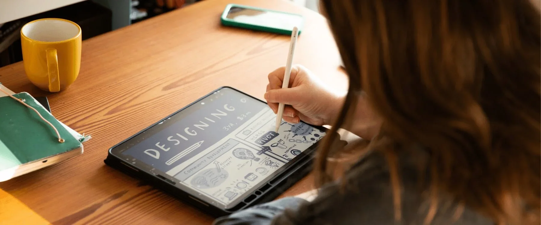 A person uses a stylus for live scribing graphics on a tablet at a wooden desk with a notebook, mug, and smartphone nearby.