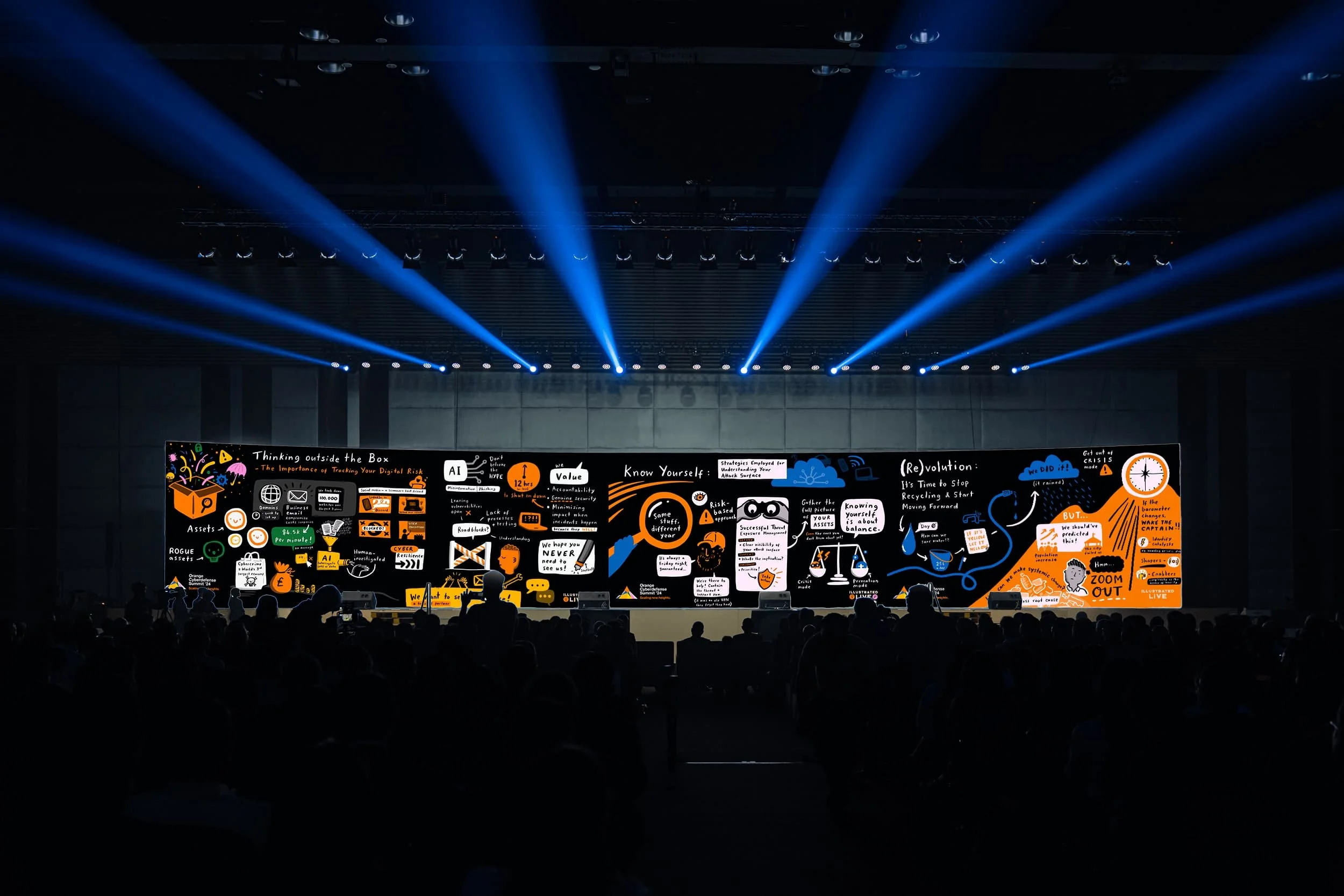 Large audience faces a brightly lit stage where live scribing and graphic illustrations are projected on a wide screen; blue spotlights above.