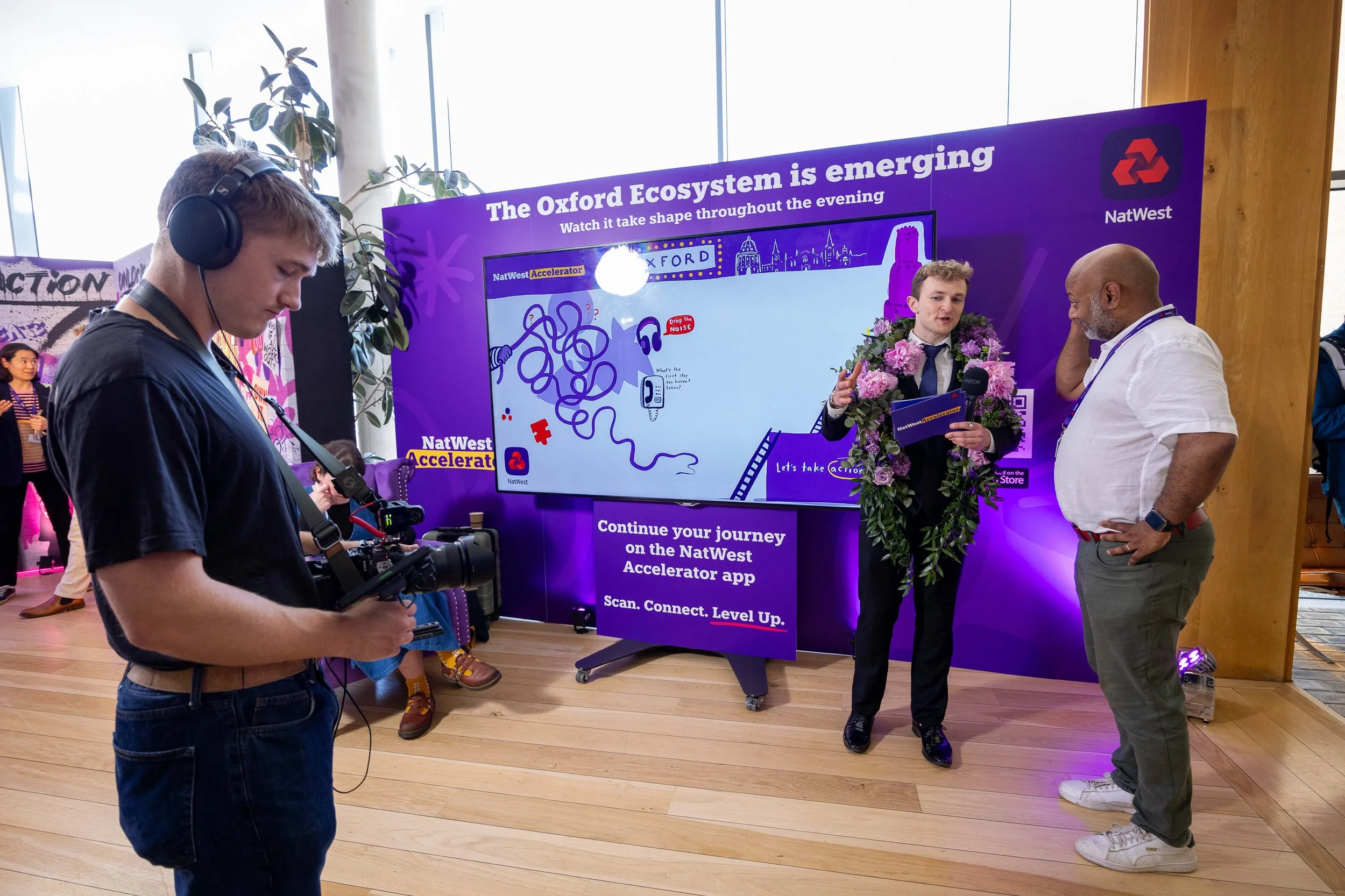 Host in a flower garland speaking into a microphone beside the live illustration screen at NatWest Accelerator.