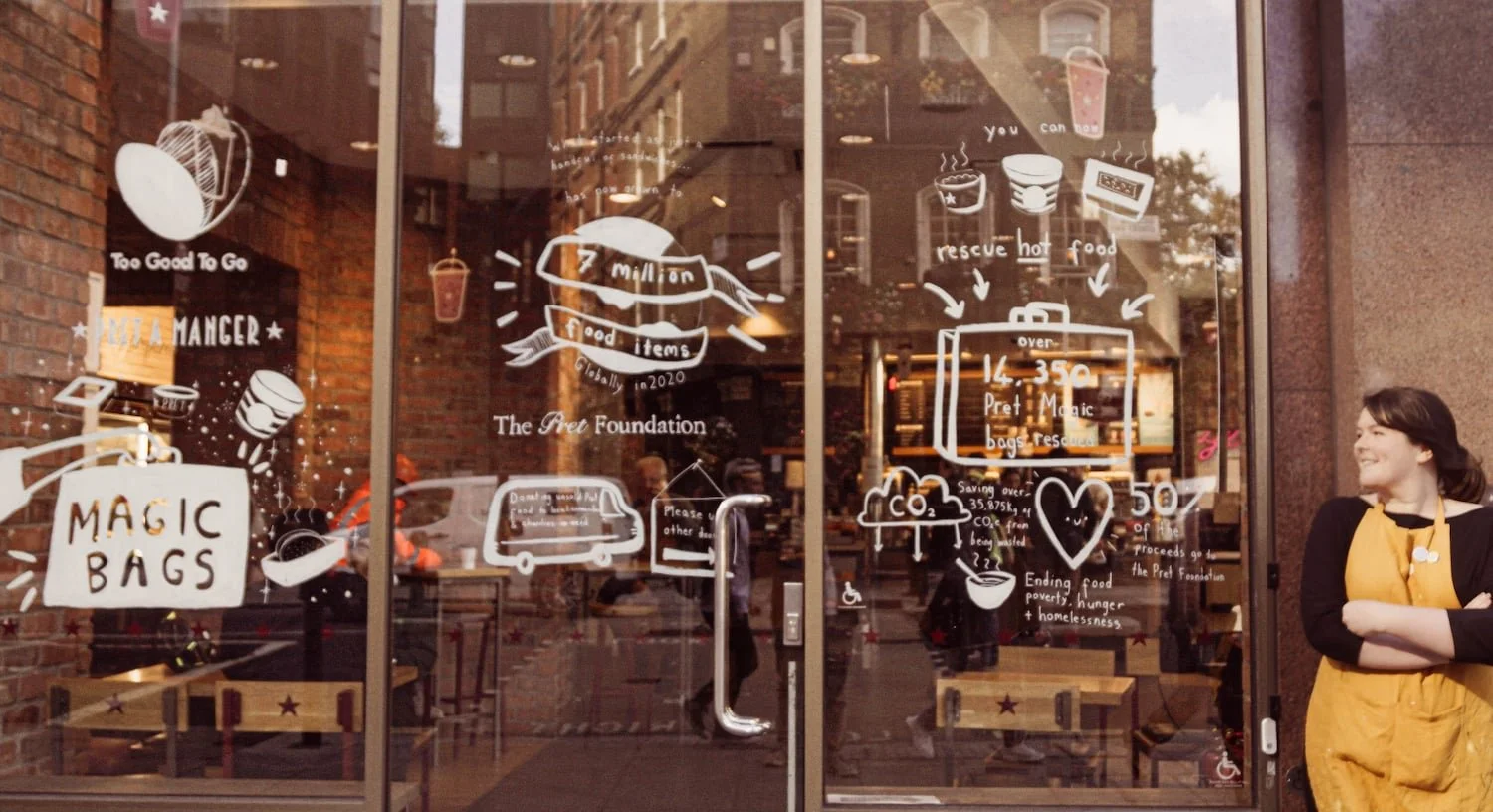 A woman in a yellow apron smiles outside a cafe with mural painting and signs about food rescue on its glass doors.
