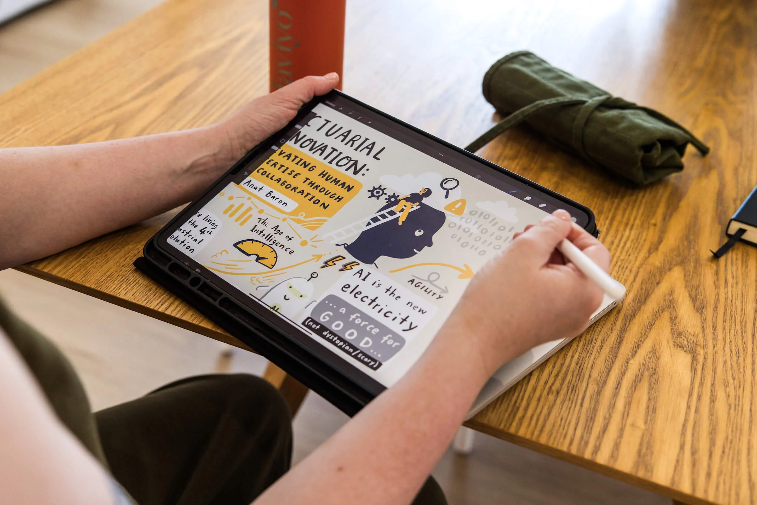Person live scribing an infographic on actuarial innovation with a tablet and stylus at a wooden table.