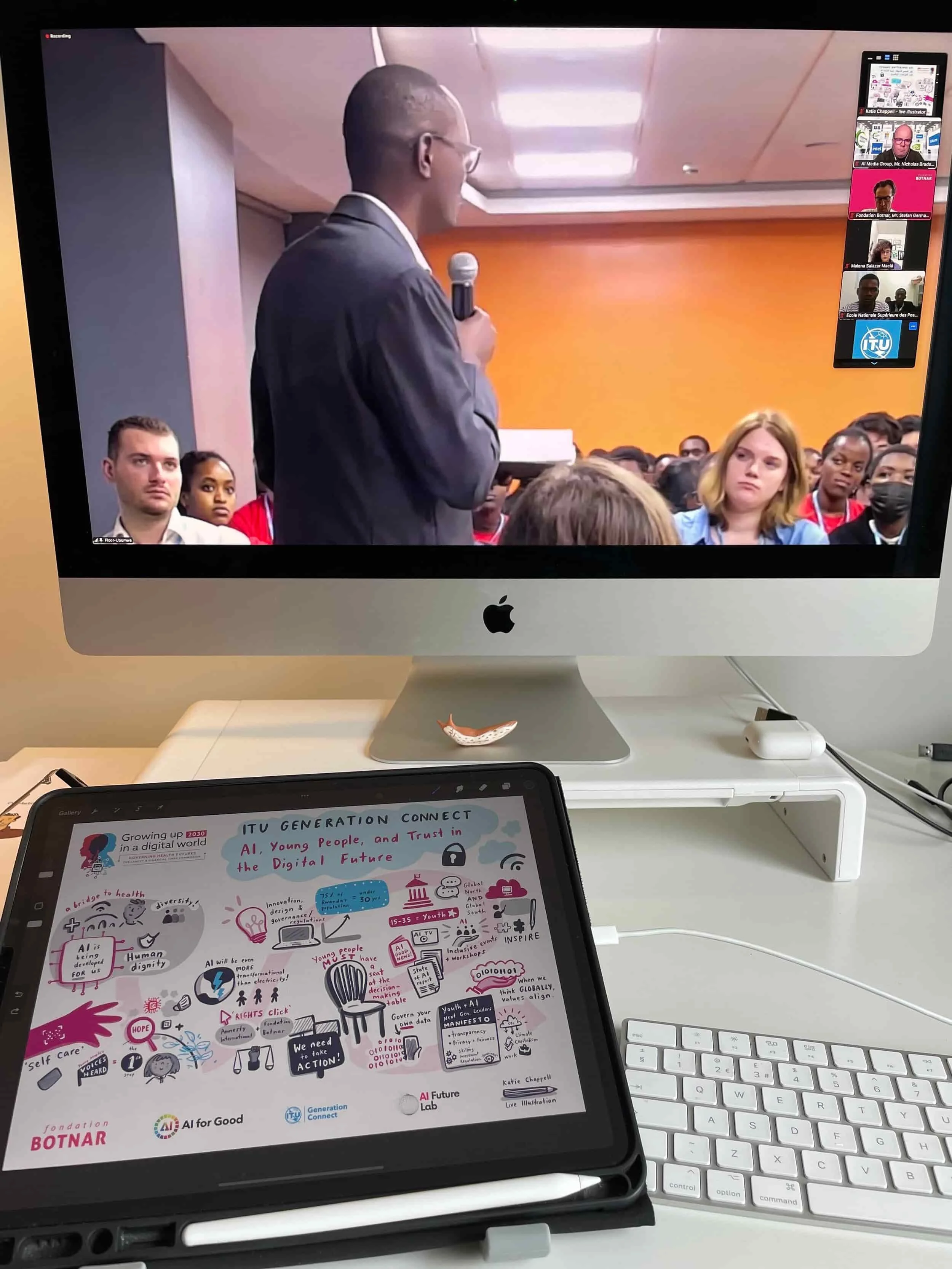 A computer screen shows a speaker addressing an audience, with live scribing notes on a tablet and a white keyboard below.