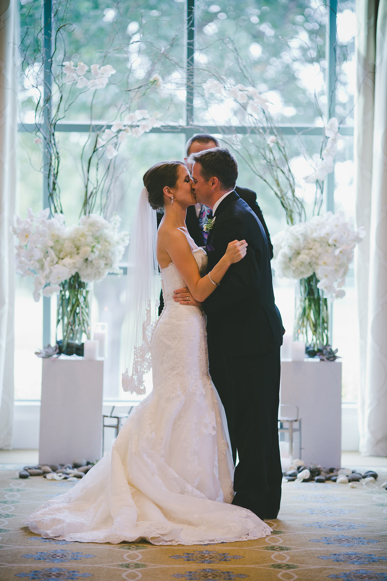 River Crest Country Club ceremony in front of large windows with a white flower and branch archway wih bride and groom kissing
