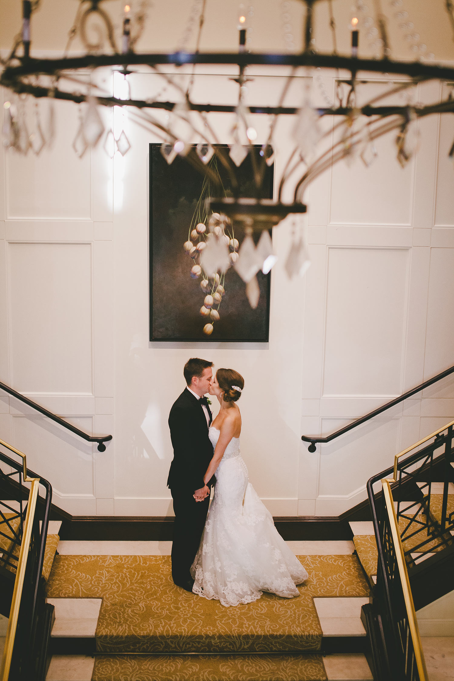 Bride and groom kissing on River Crest Country Club stairway behind crystal chandelier