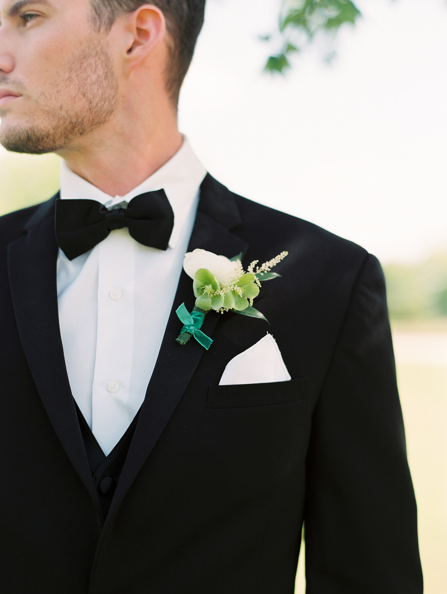 Groom in black tuxedo and black bow tie with a white boutonniere and green ribbon