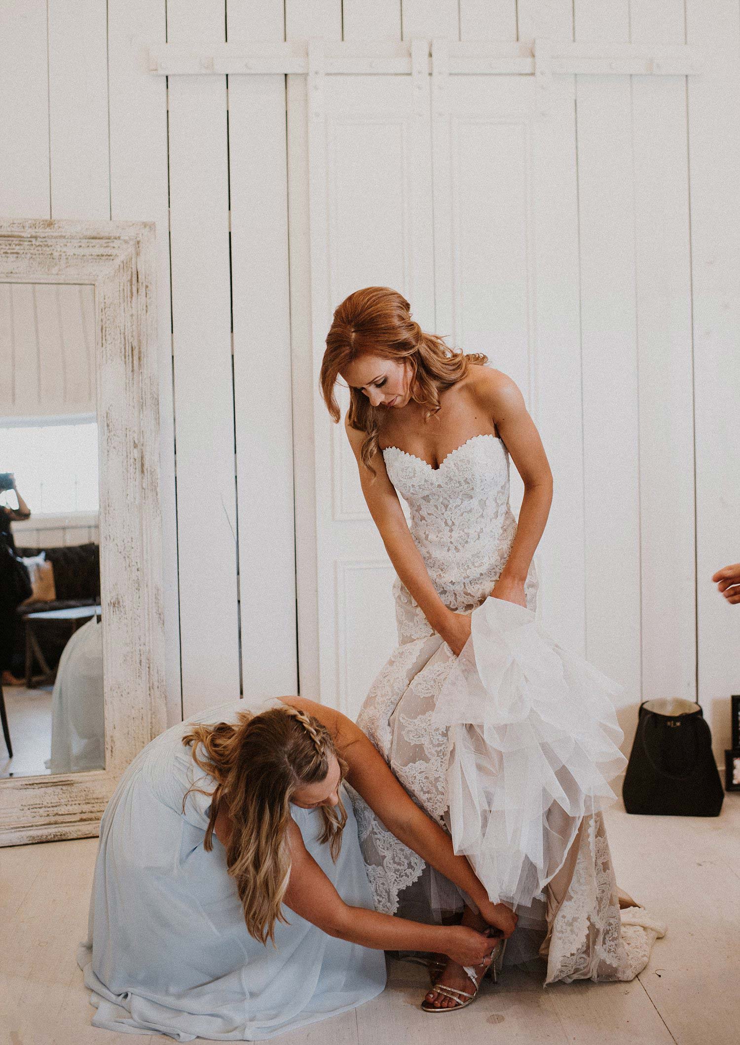 Bride putting on lace wedding dress with bridesmaid in long dusty blue bridesmaid dress at White Sparrow Barn