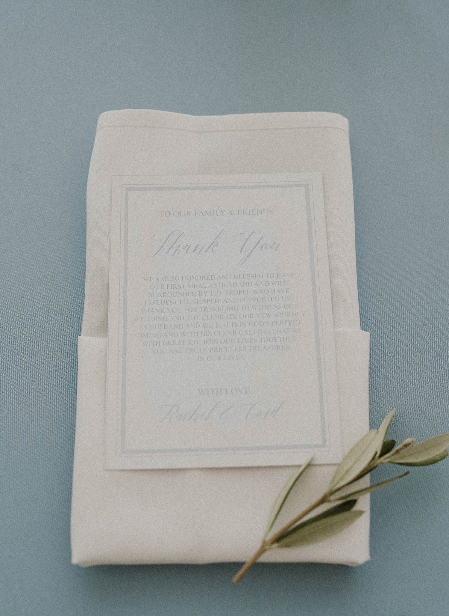 Menu on folded napking with olive branch on top of light blue linen