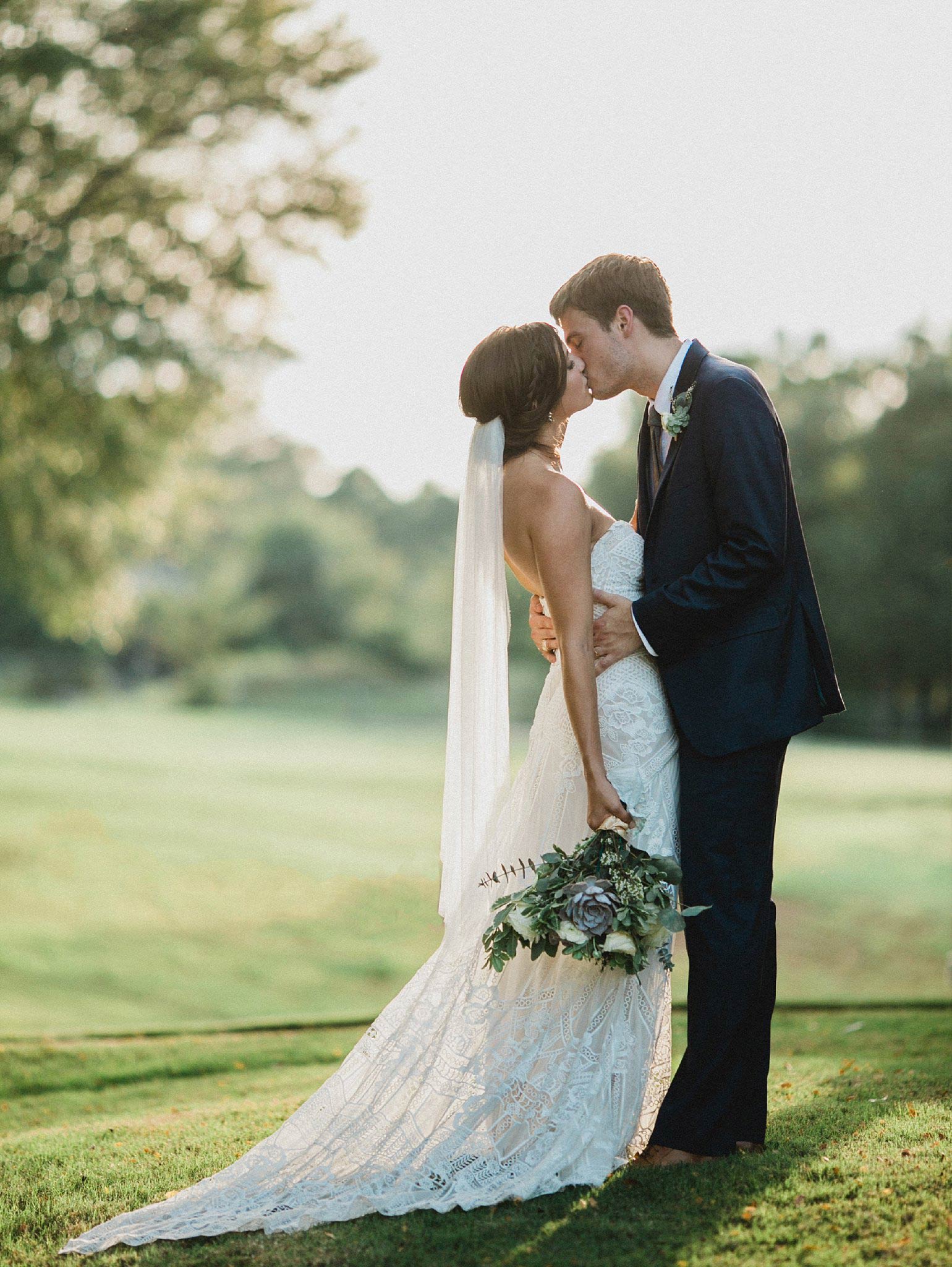 Bride wearing a lace boho dress kissing the groom while holding a greenery and succulent bouquet 