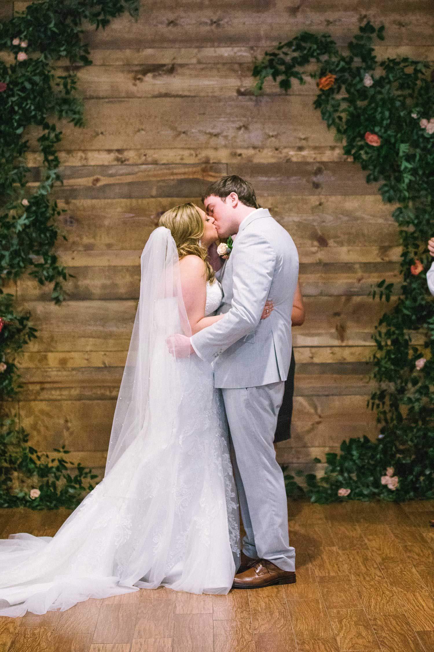 Bride and groom kissing infront of greenery archway against wood wall