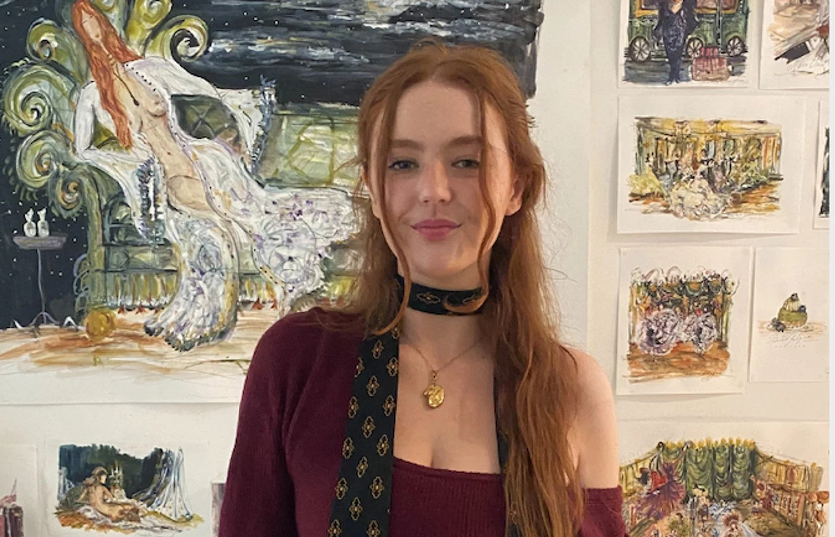 Rachelle Cunningham with long red hair standing in front of a gallery wall featuring colorful, expressive artworks. She is wearing a burgundy off-the-shoulder top, a black scarf with a gold pattern, and a gold pendant necklace.