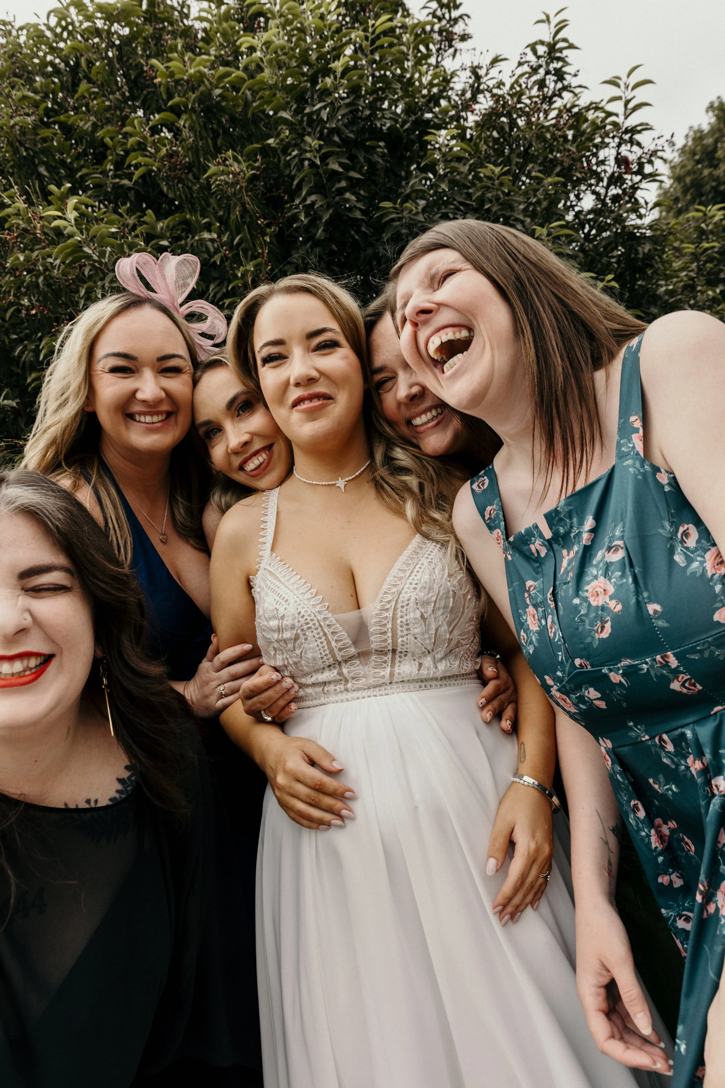 Group of women smiling and enjoying a fun moment outdoors, dressed in colorful and elegant clothing, with a lush green background.