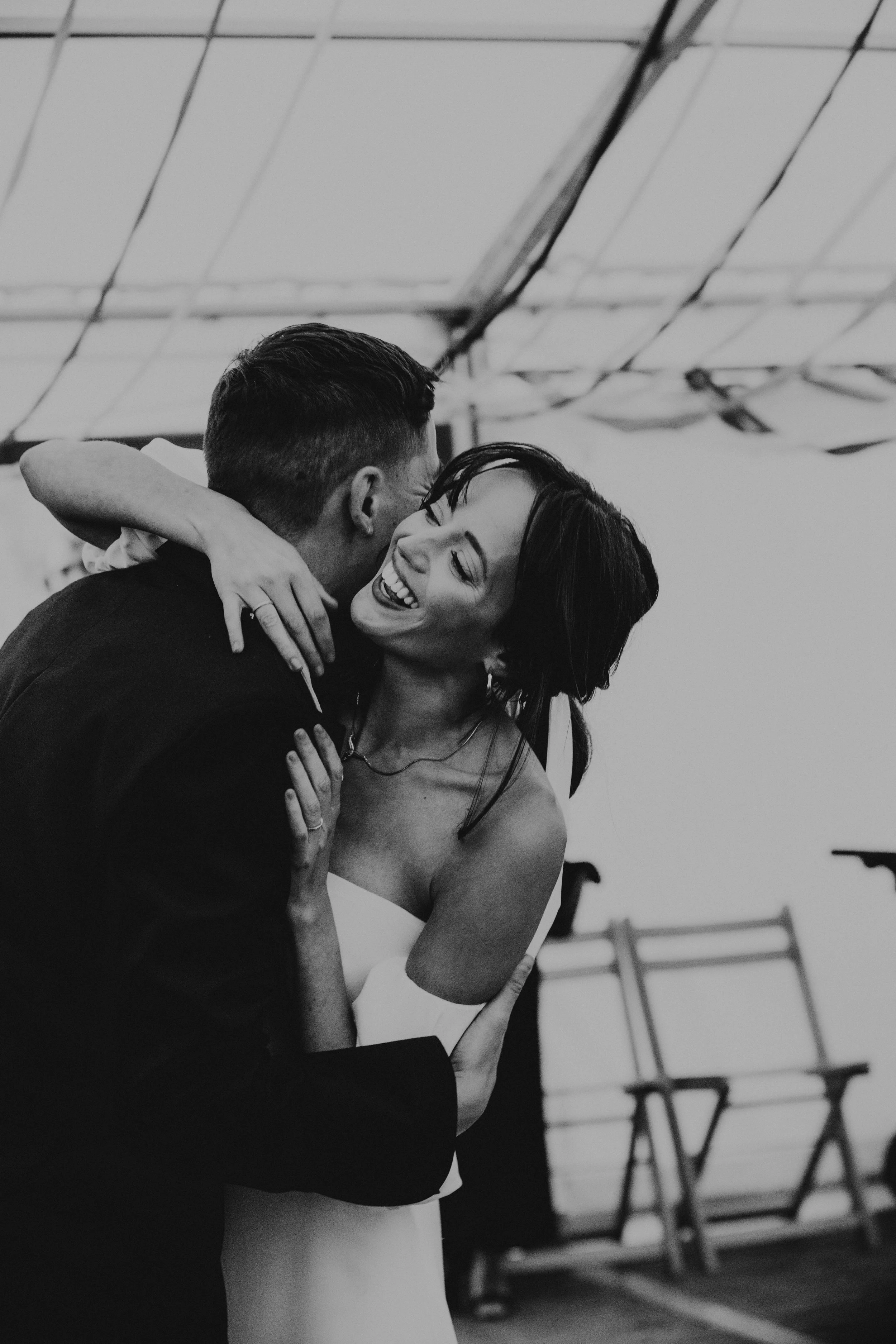 A man in a suit and a woman in a dress are dancing closely and smiling at each other indoors, with chairs in the background.