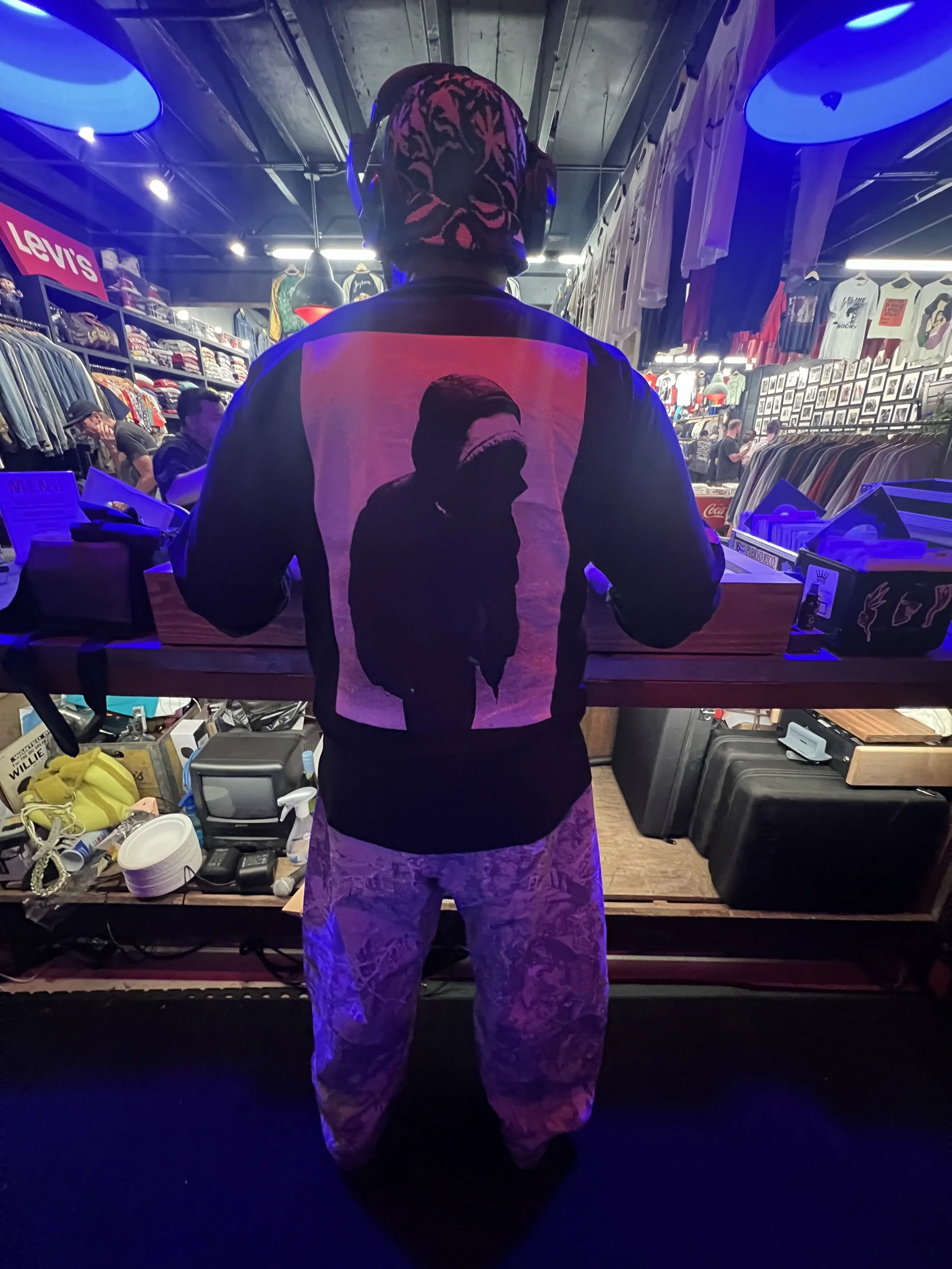 Person wearing a hoodie with a shadowed face image on the back, standing behind a counter in a store with clothing and framed pictures on the wall, illuminated by blue and purple lights.