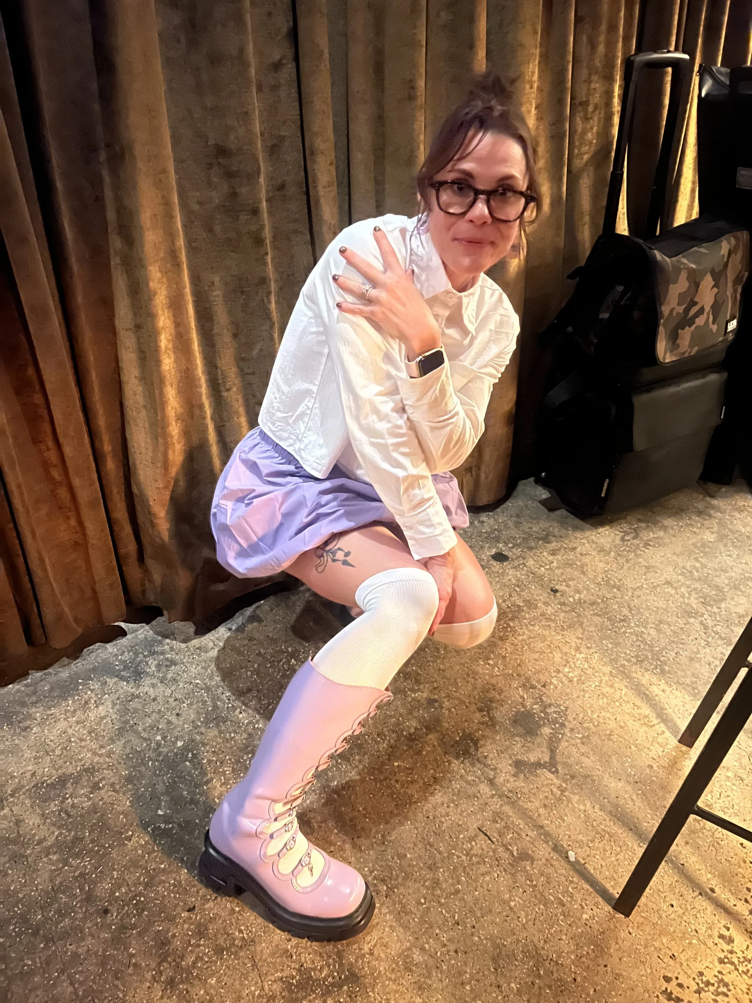A woman dressed in a white shirt, pink skirt, and pink knee-high boots with chunky soles, sitting on her heels in front of a wooden wall, with her hand on her shoulder, wearing glasses and a smartwatch.