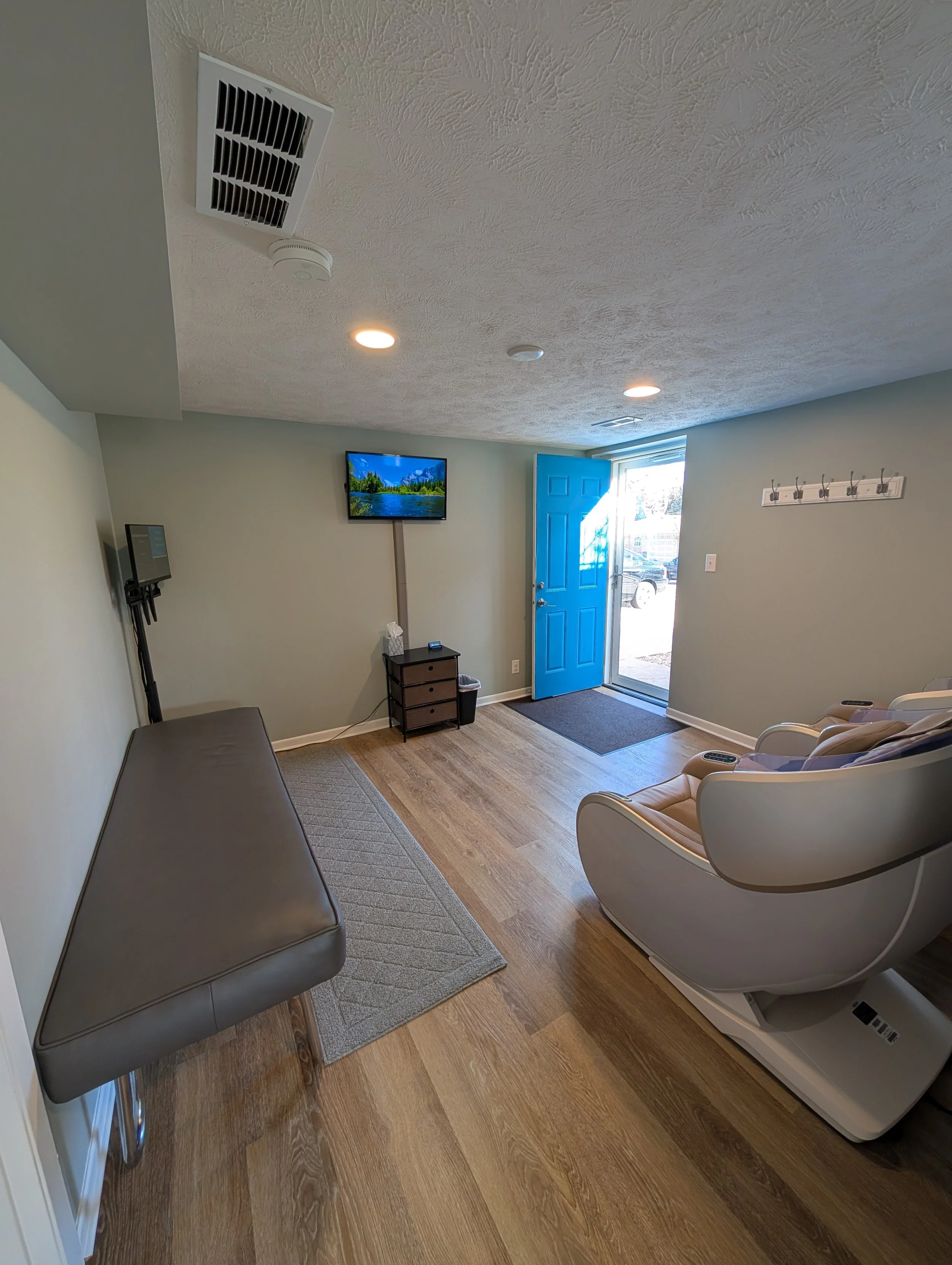 Waiting room with massage chairs, wall-mounted TV, small black side table, coat hooks, and open blue front door letting in sunlight.