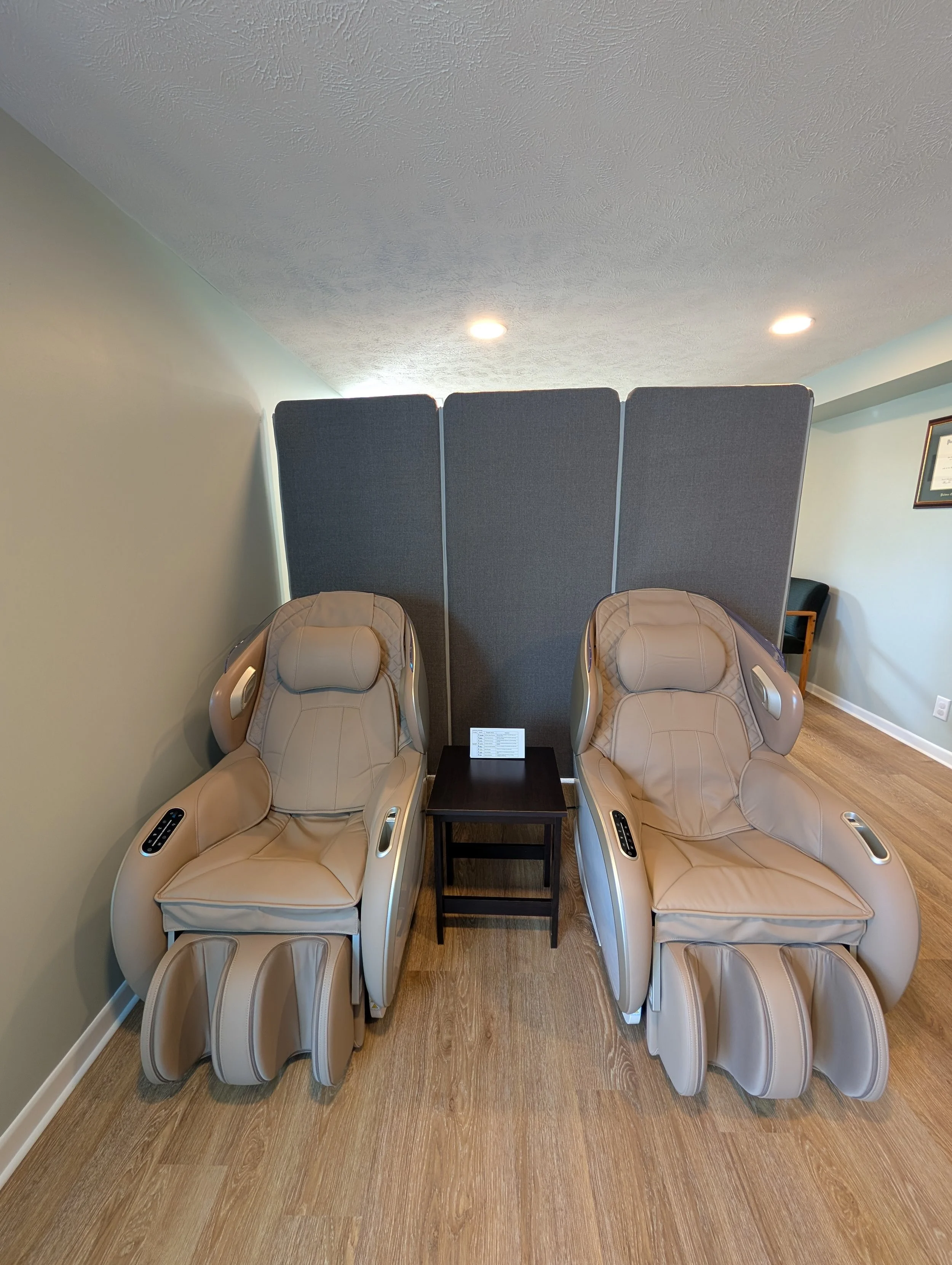 Two beige massage chairs facing forward with foot rollers, placed side by side, with a small black table between them, in a room with wood flooring and a privacy screen behind the chairs.