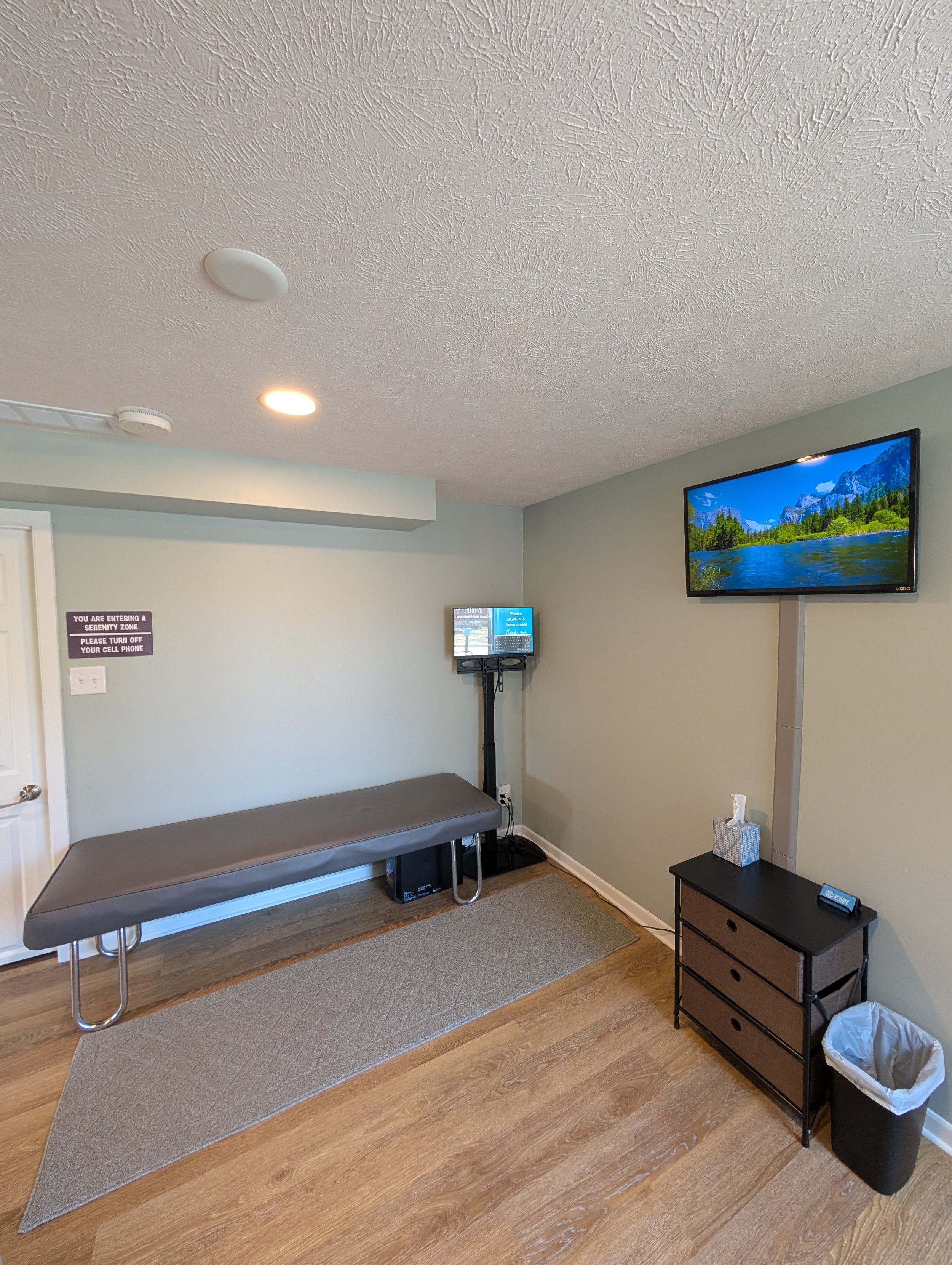 Interior of a medical examination room with a padded examination table, a wall-mounted TV displaying a nature scene, and a small black dresser with tissues and a trash can nearby.