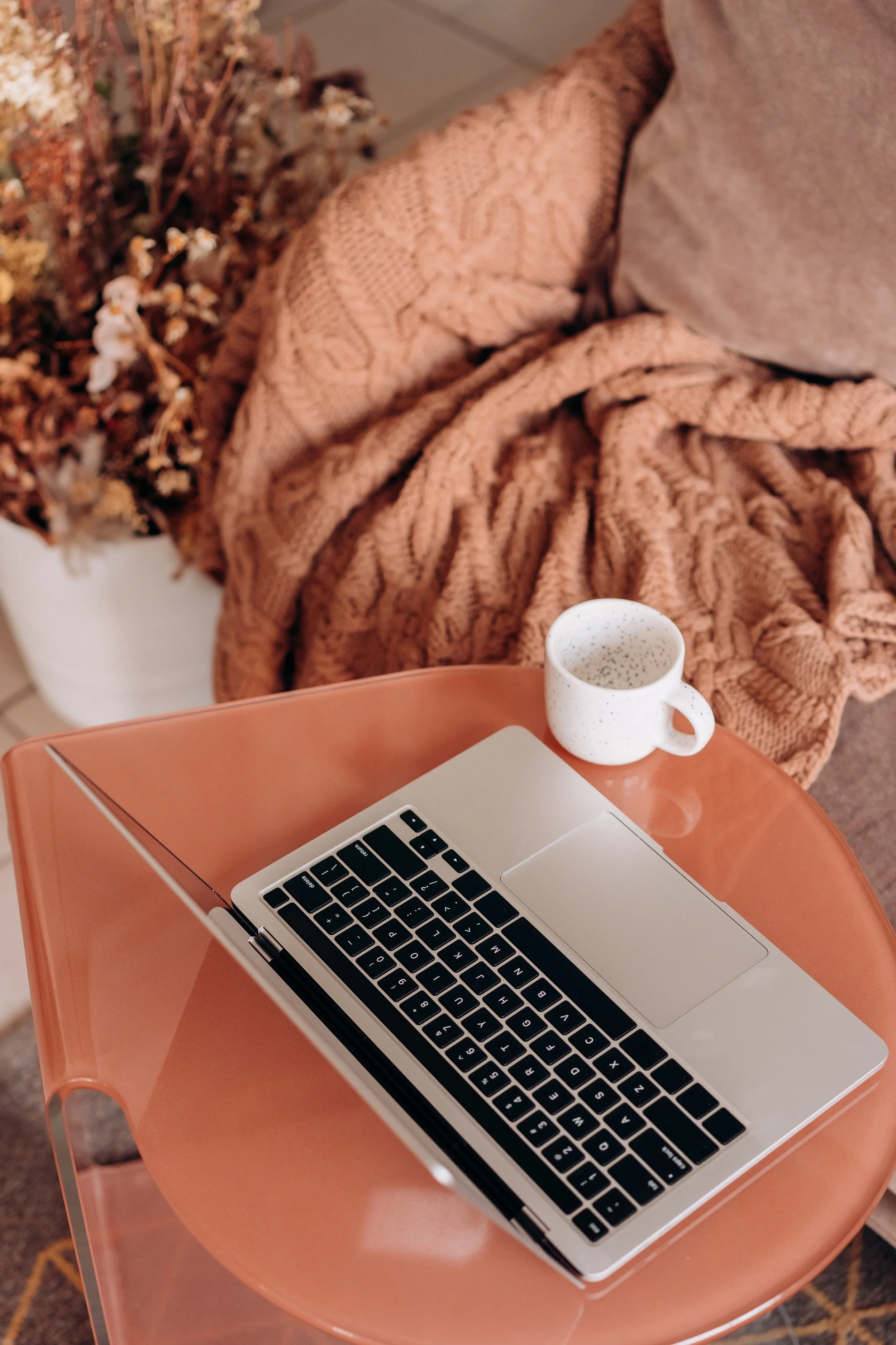 A laptop, a white speckled mug, and a pinkish-orange tray on a small table. In the background, a sofa with a beige pillow and a chunky pink knitted blanket, and a pot with dried flowers.