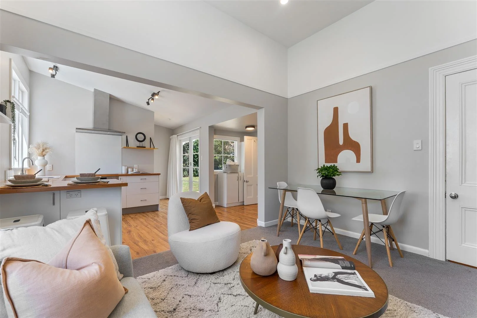 Modern open-concept living room and kitchen with white walls, wood flooring, and minimalistic decor including a white couch, round white chair with pillow, and a glass dining table with white chairs. Decor includes vases, artwork, and plants.