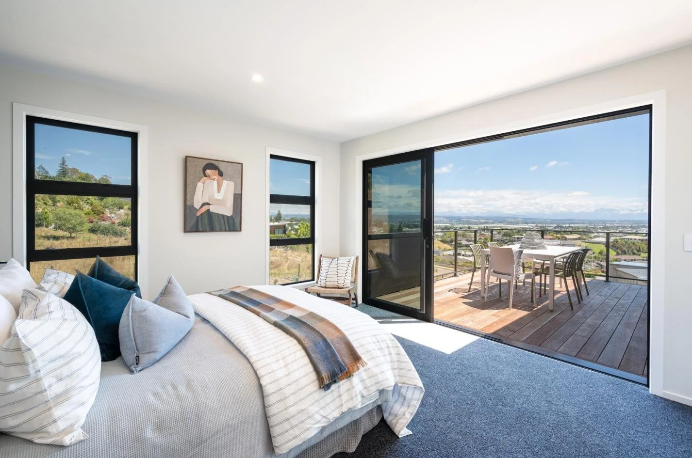 Bedroom with white walls, large glass sliding door leading to a balcony with outdoor dining, a bed with pillows and a blanket, and artwork on the wall.