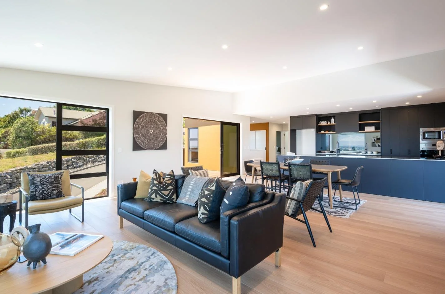 Open-concept living room and kitchen with modern furniture, black sofa with patterned pillows, dining table, black cabinets, and large windows showing outdoor greenery.