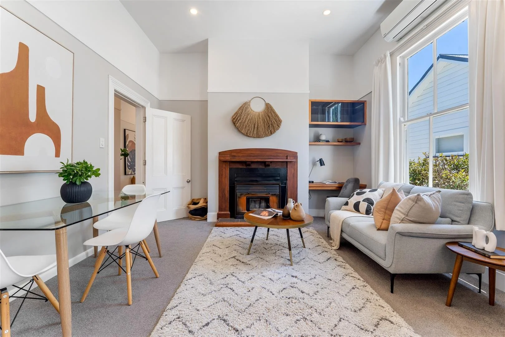 Living room with large window, white sofa, wooden coffee table, fireplace with wooden mantel, wall art, and a small desk area, decorated in neutral tones and modern style.