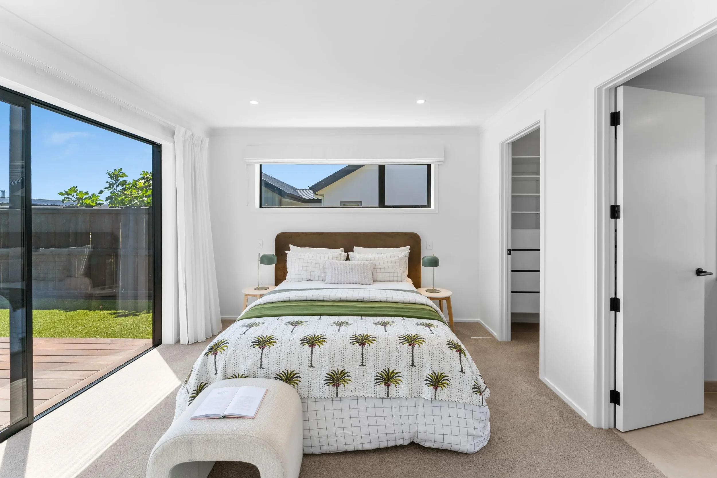 A bright bedroom with white walls and a large bed with a palm tree patterned duvet, two bedside tables with green lamps, and sliding glass door leading to an outdoor deck and yard.