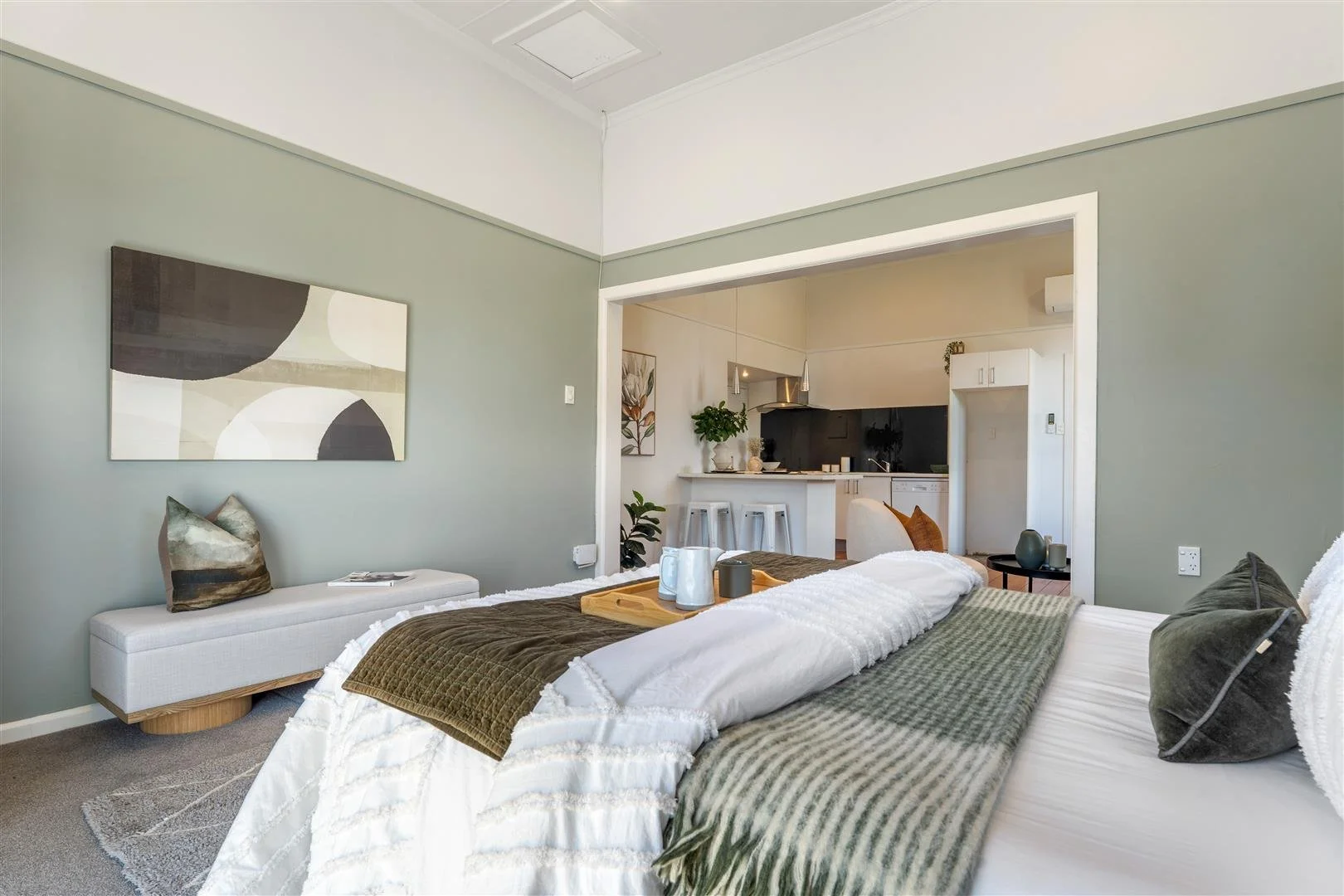 A cozy bedroom with a bed covered in white, brown, and green blankets, decorative pillows, and a tray with mugs on the bed. There is a white bench with a pillow and magazine, a wall art piece above, and an open view into a kitchen with bar stools, pl